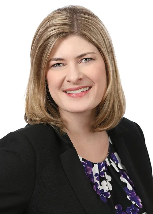 Professional woman with shoulder-length blonde hair smiling, wearing a black blazer over a floral blouse, against a white background.