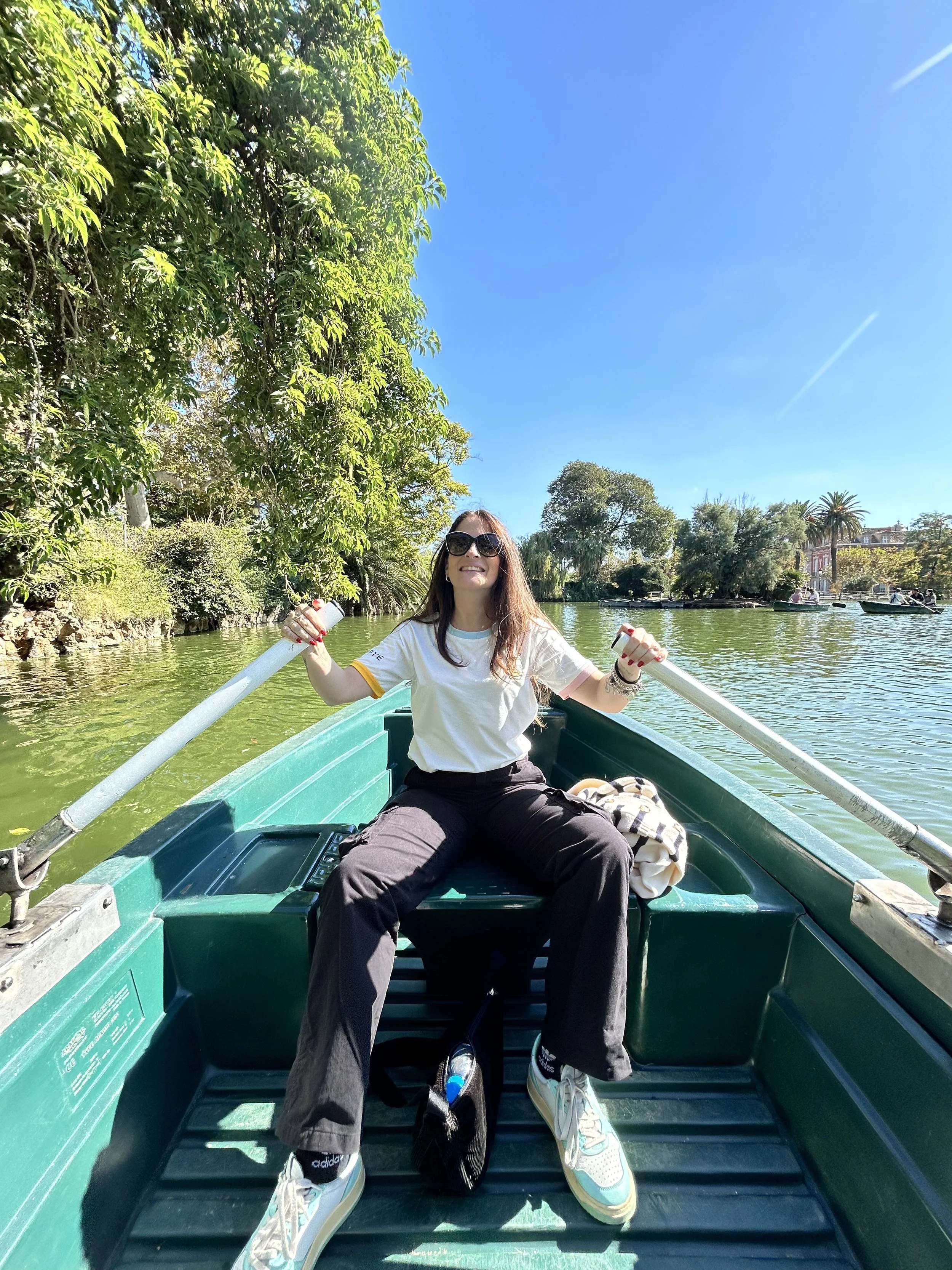 Una donna seduta su una barca con oars in una zona tranquilla con alberi e acqua, sotto un cielo azzurro soleggiato.