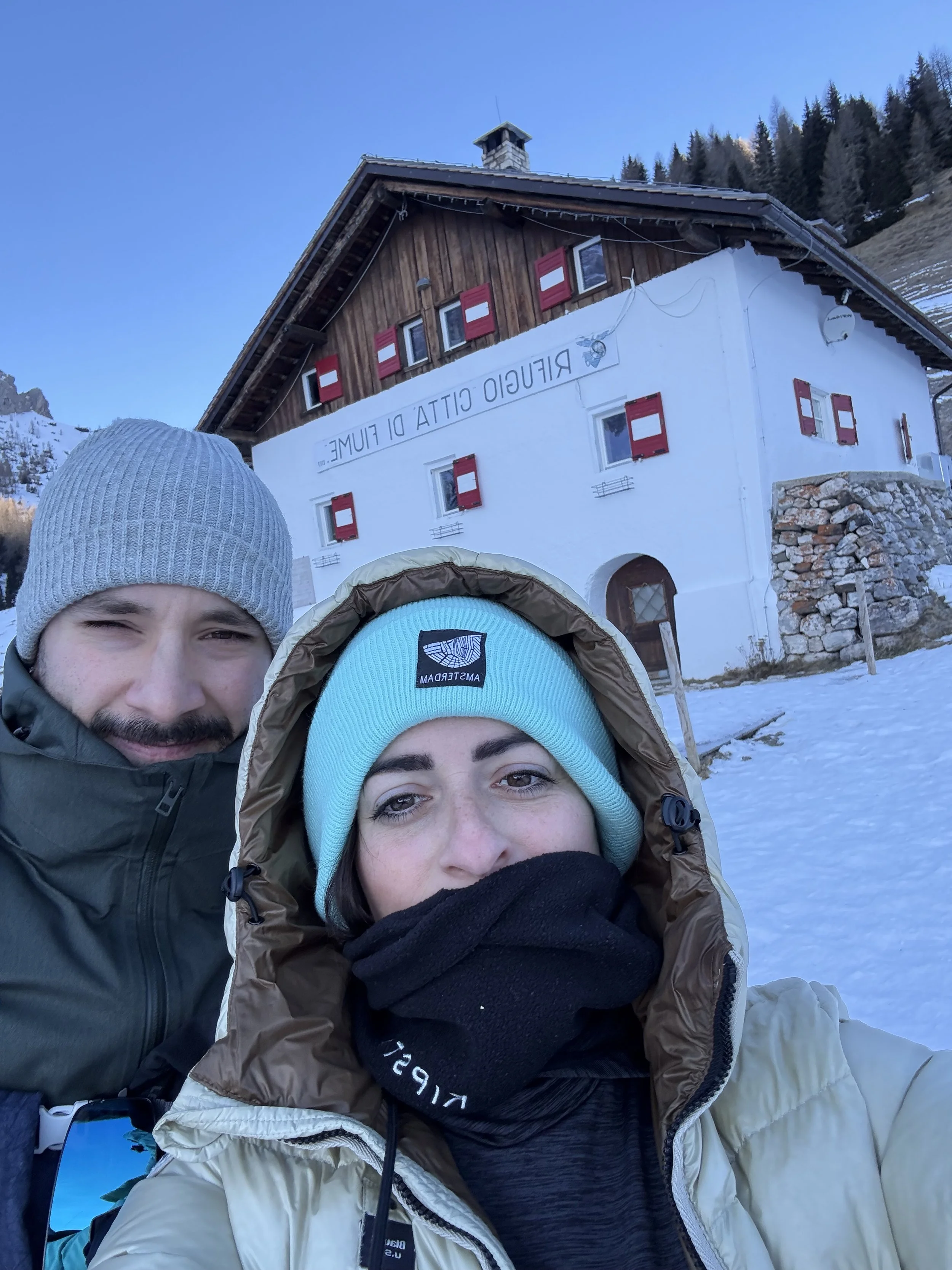 Due persone vestite in abbigliamento da neve si trovano all'aperto con una baita in neve alle spalle. La baita è bianca con tetto in legno e scritte in italiano, circondata da alberi e montagne.