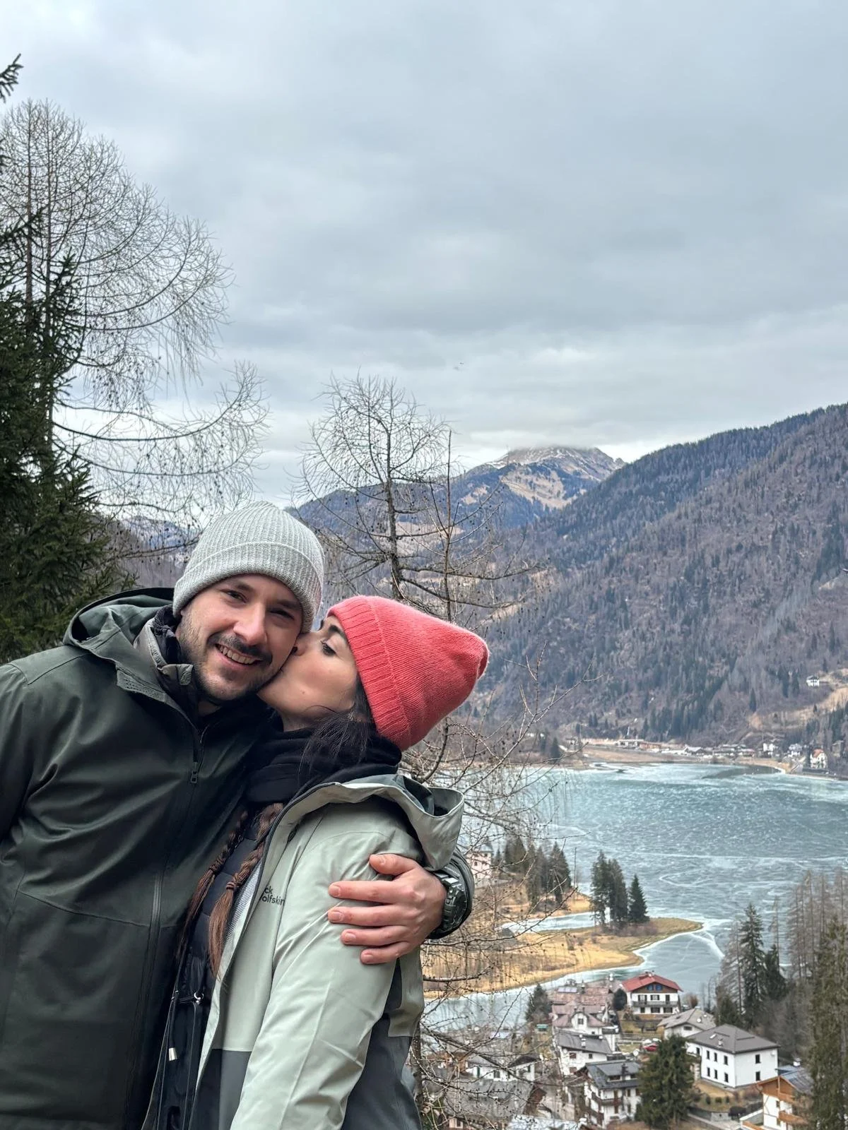 Una coppia sorridente abbracciata con uno sfondo di montagne, un lago e un villaggio in un paesaggio invernale