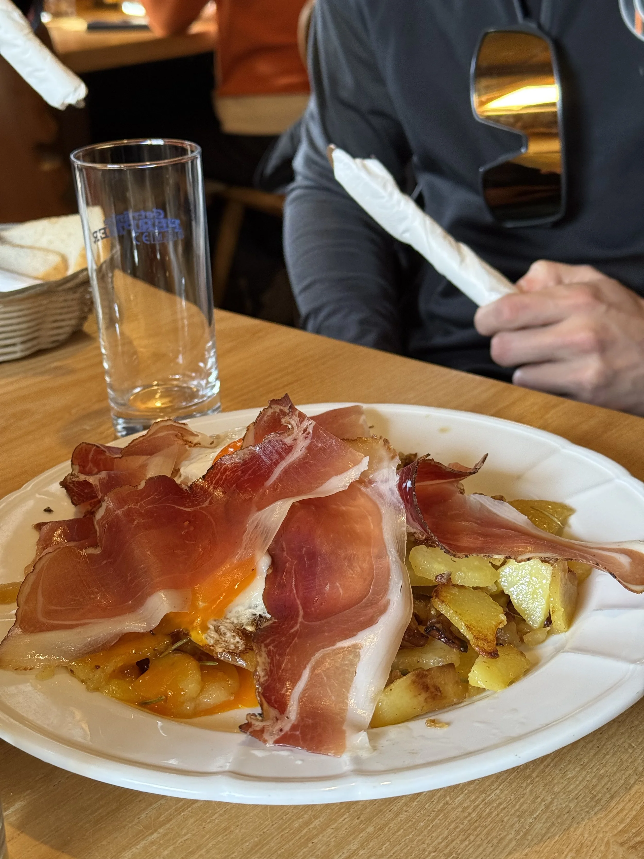 Piatto di patate arrosto con prosciutto crudo e un uovo in camicia, in un piatto di plastica bianca su un tavolo di legno