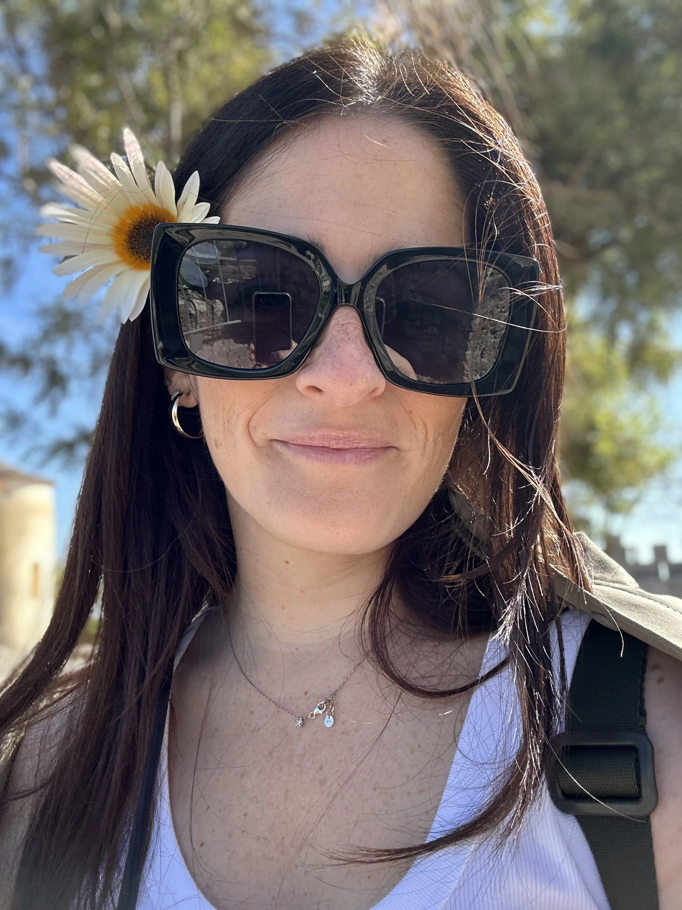 Una donna con capelli lunghi, occhiali da sole grandi e una margherita infilata tra i capelli, sorridente, in una giornata di sole all'aperto con albero e cielo azzurro sullo sfondo.