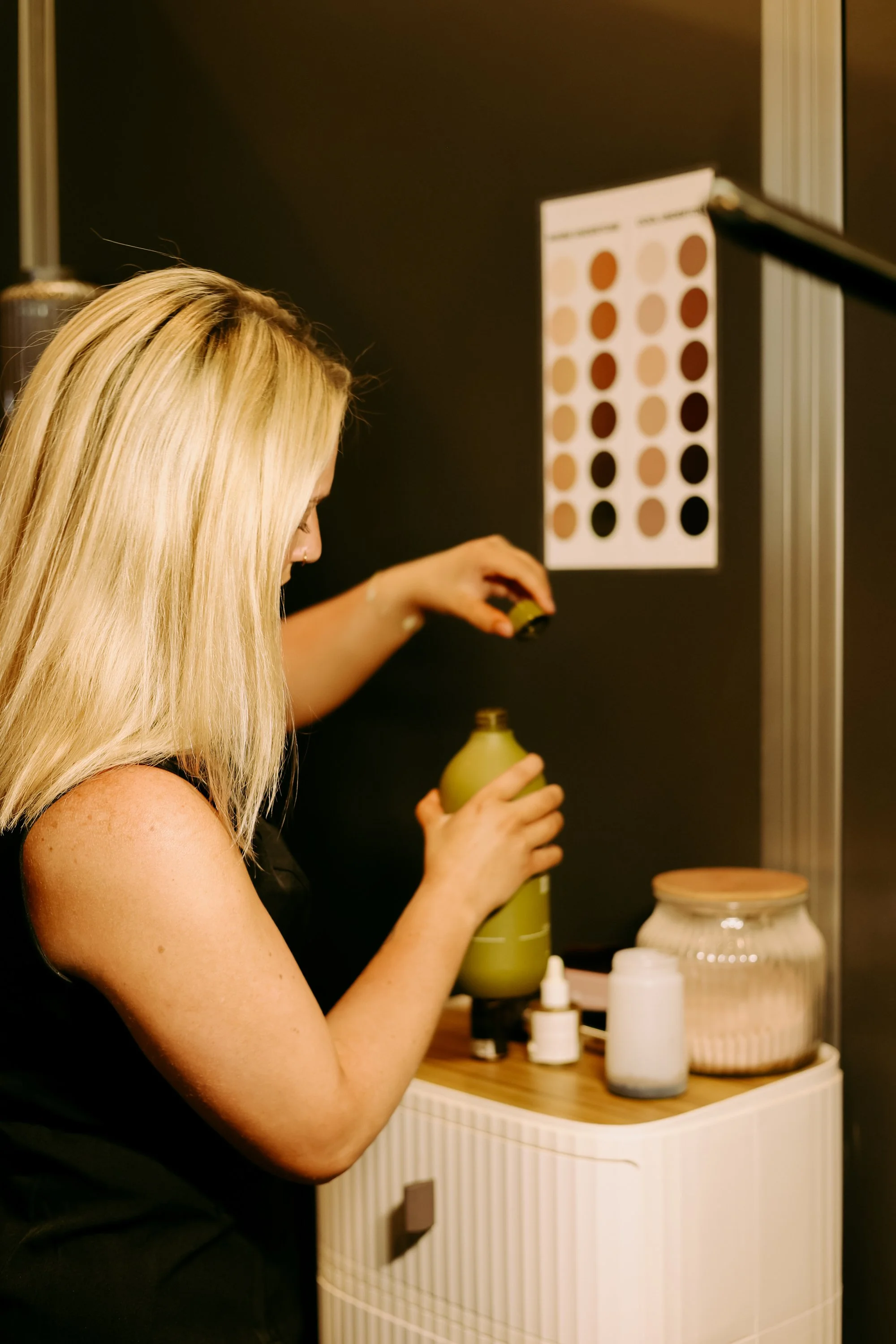 A woman with blonde hair preparing a green beverage or medicine in a bottle in a modern kitchen or lab setting.