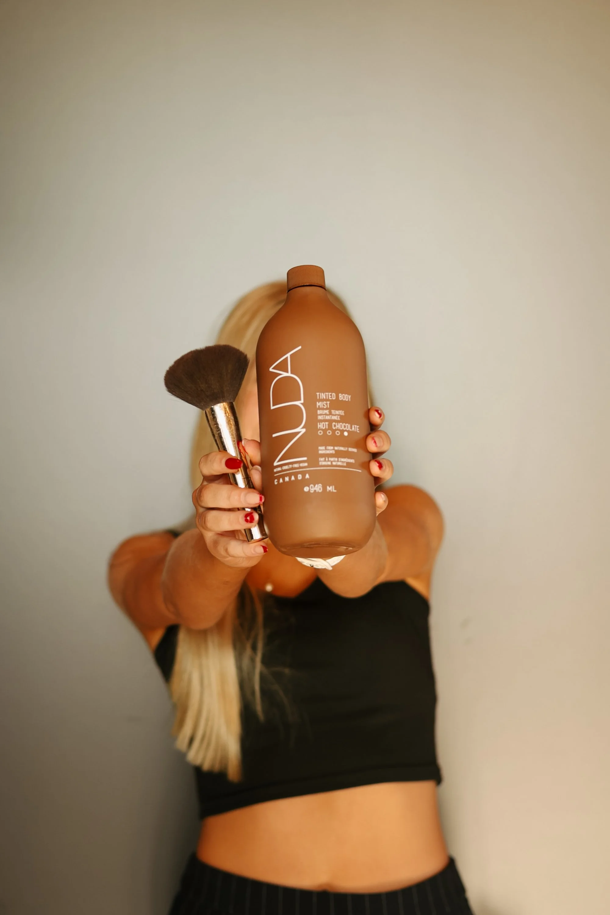 Person holding up a tan bottle of INNODA Tinted Body Mist with a makeup brush, facing the camera. The person is wearing a black crop top and has long blonde hair.