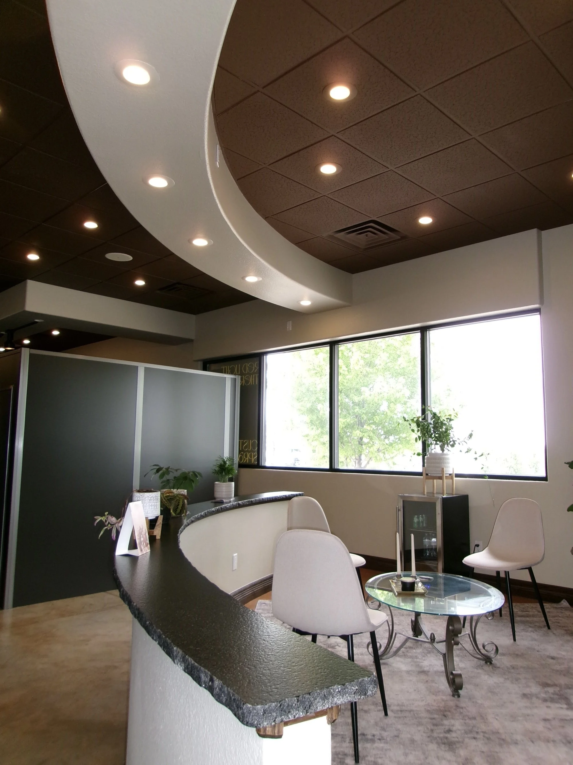Modern waiting area with curved reception desk, white chairs, glass coffee table, and large window with greenery outside.