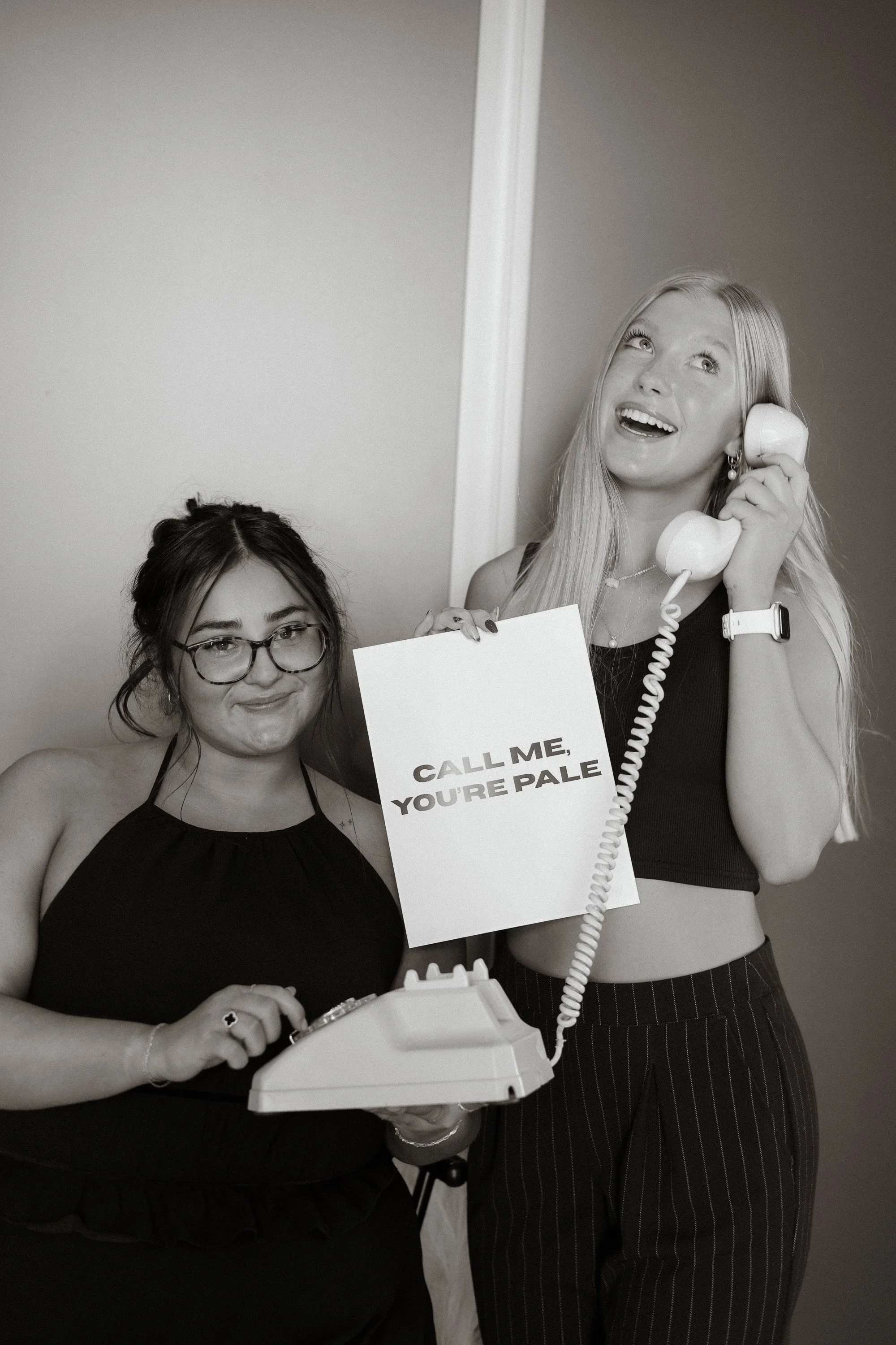 Two women are standing together, one holding a vintage telephone receiver to her ear and the other holding a sign that reads, "CALL ME, YOU'RE PALE." The woman on the left has dark hair, glasses, and is wearing a sleeveless top, while the woman on th