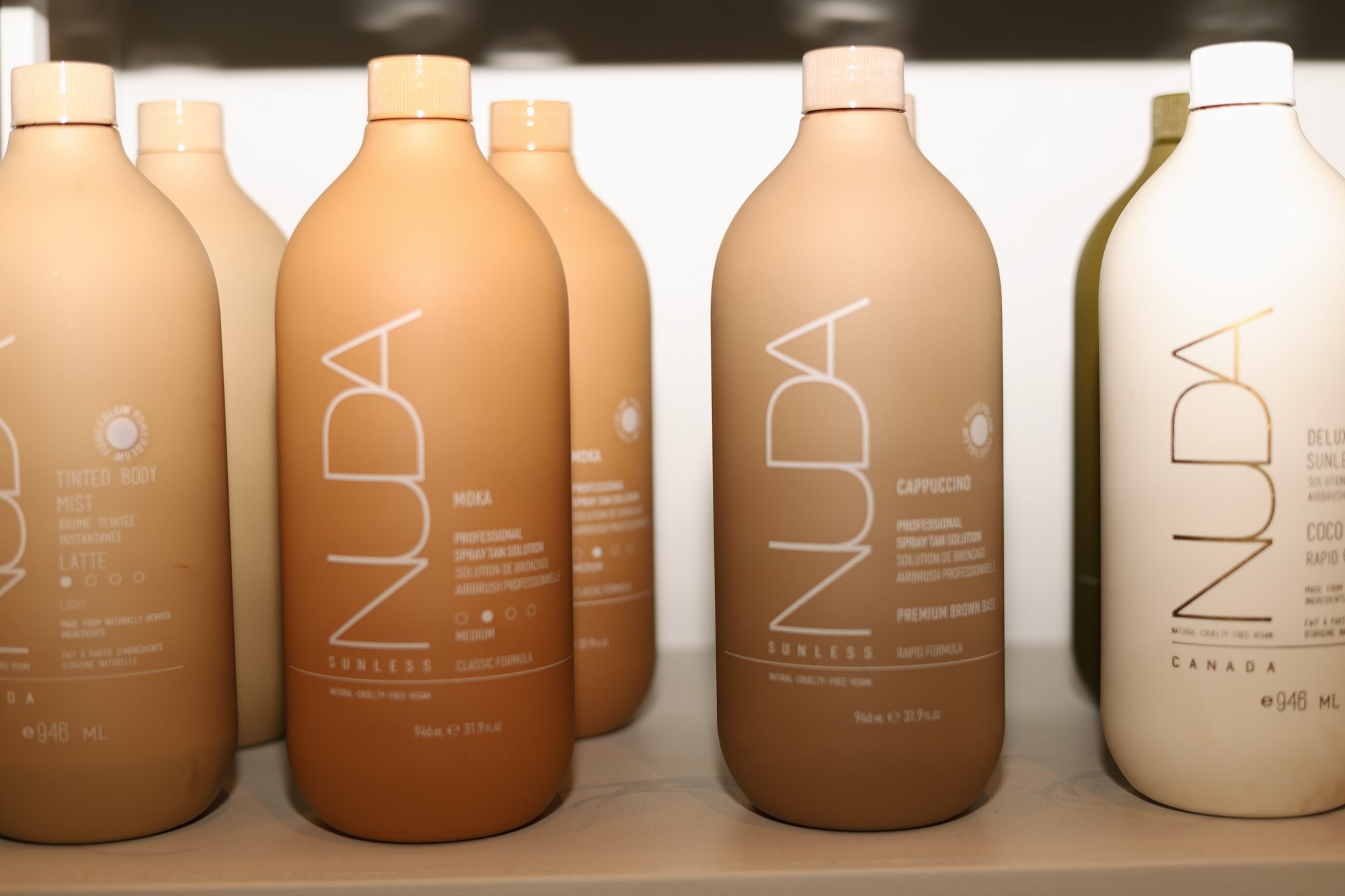 Bottles of ZUDA sunless tanning products on a shelf, including different formulas such as latte, mocha, cappuccino, and coconut, in beige, brown, and white bottles.