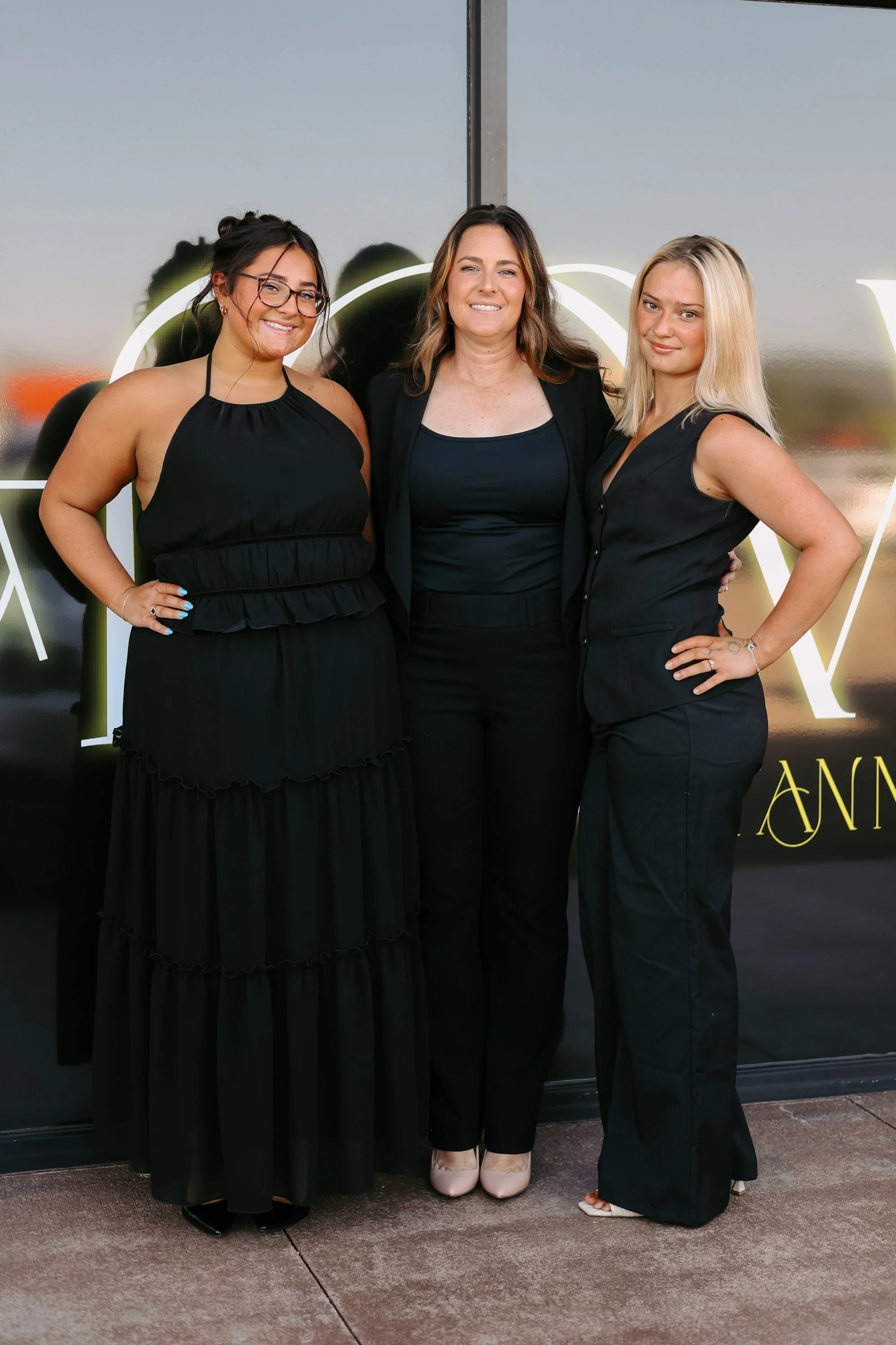 Three women posing together for a photo, dressed in black and standing in front of a glass wall with illuminated art.