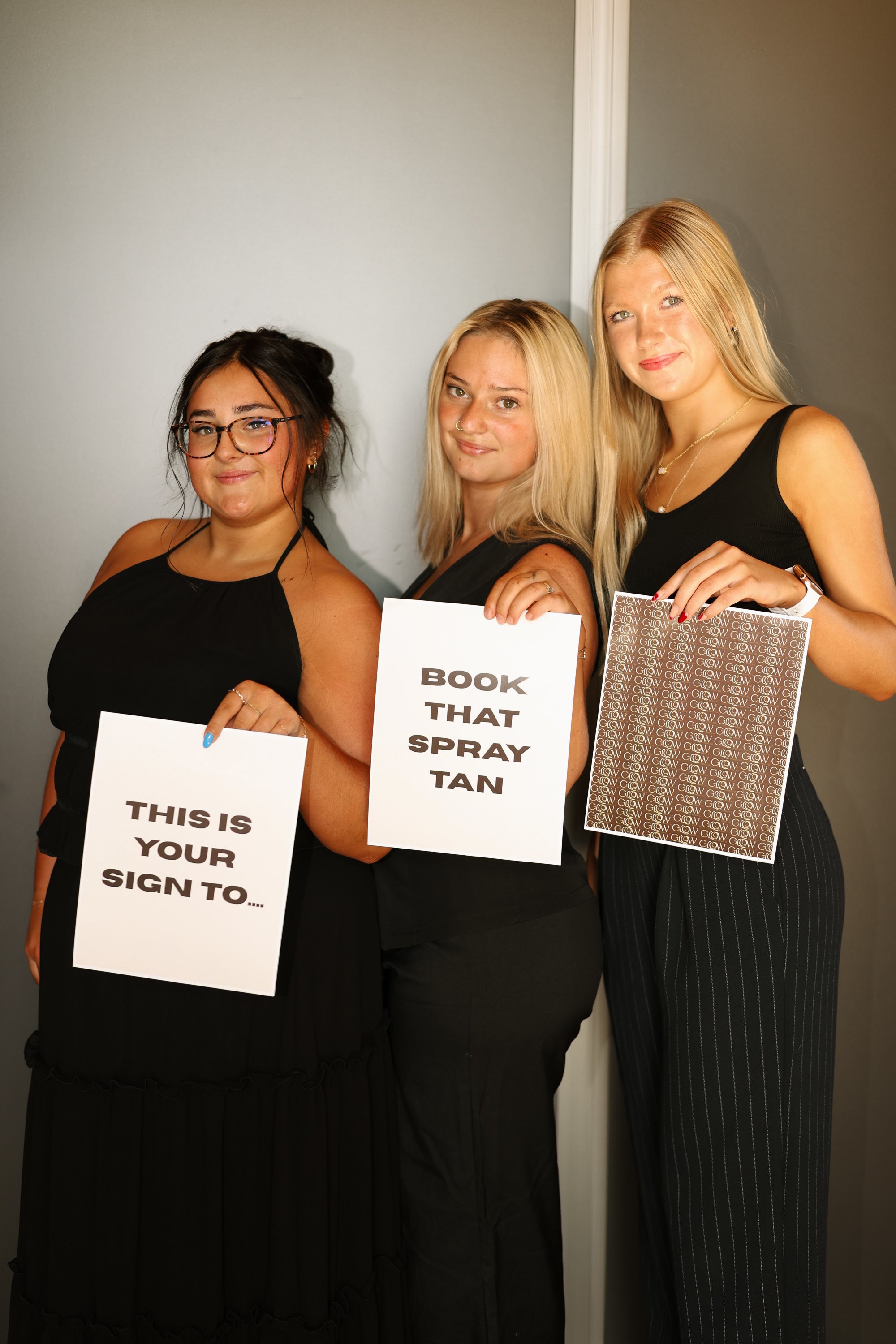Three women standing side by side indoors, holding signs with humorous messages about nail polish and hair spray, dressed in black and business casual attire.
