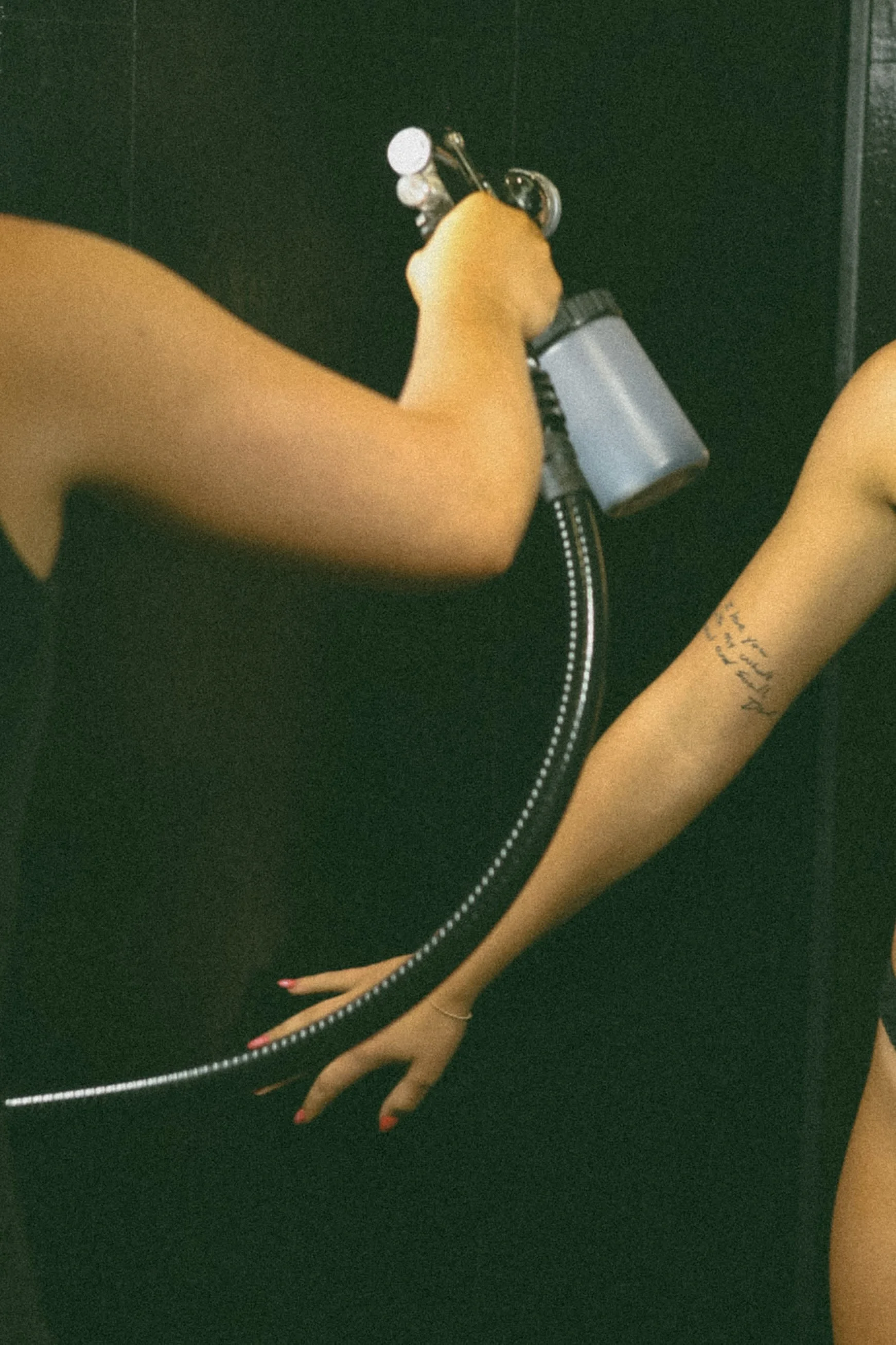 Close-up of a person spraying perfume from a bottle in a black outfit with a silver zipper, with part of another person visible, including an arm with a tattoo.