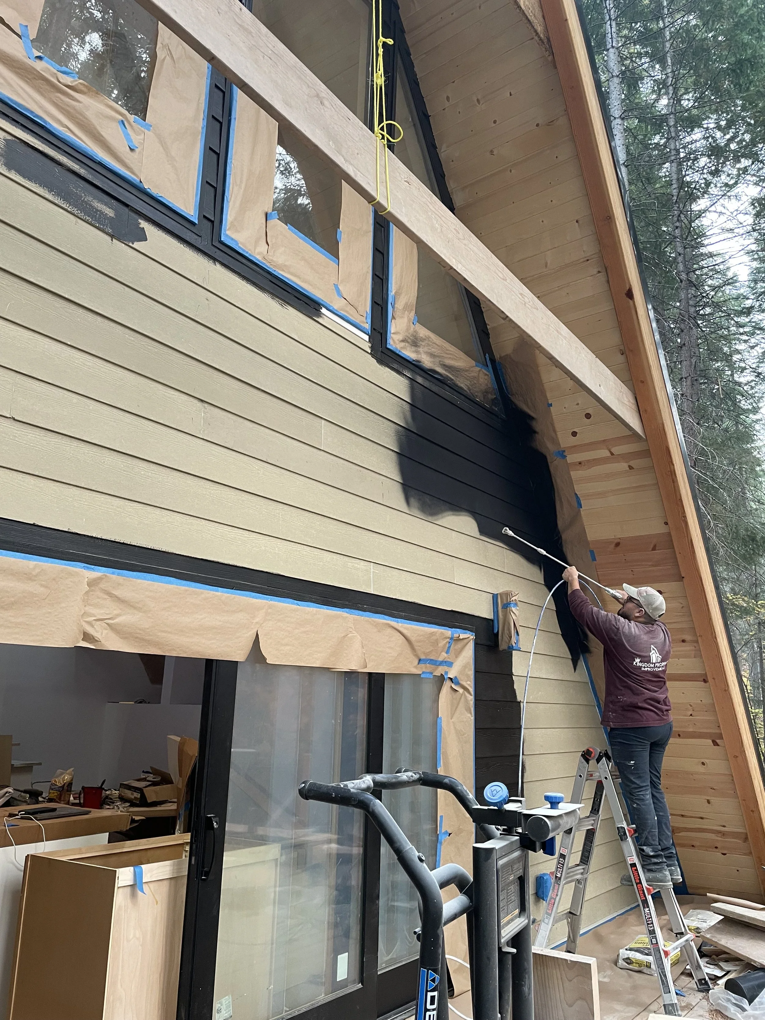 A person painting the exterior of a house black while standing on a ladder.