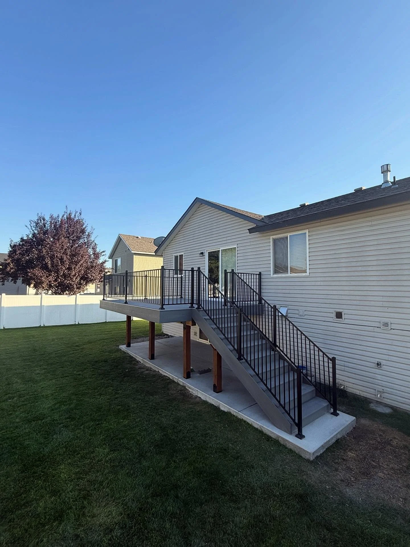Backyard view of a house with a new elevated deck, stairs, and black metal railing under a clear blue sky.