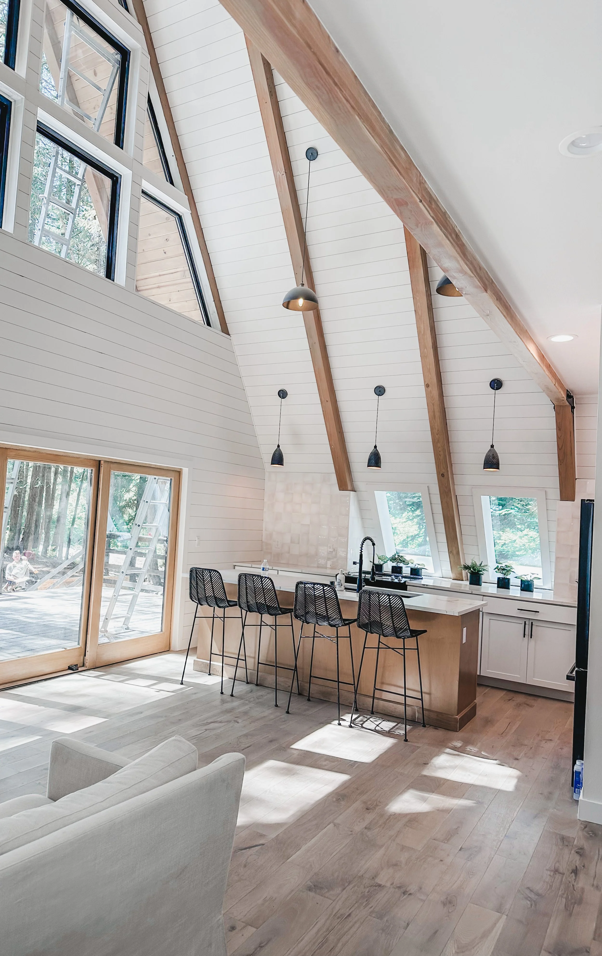 A modern kitchen in a bright open-plan living space with high vaulted ceiling, exposed wooden beams, large windows, and sliding glass door. The kitchen features a white island with four black bar stools, white cabinetry, and a black faucet. Potted pl
