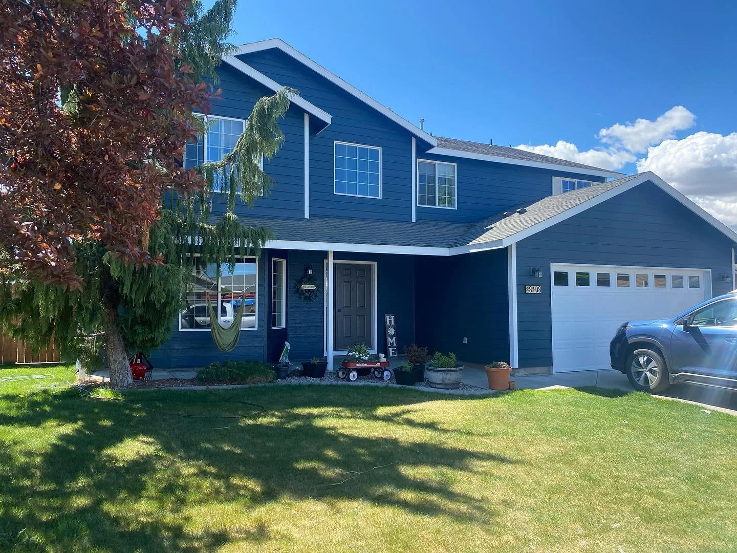 A blue two-story house with a garage, a front lawn, and a large tree. The house has white trim, windows, and a front door with a wreath. A small hammock hangs beside the house, and potted plants decorate the porch area. A silver car is parked in the driveway under a partly cloudy sky.