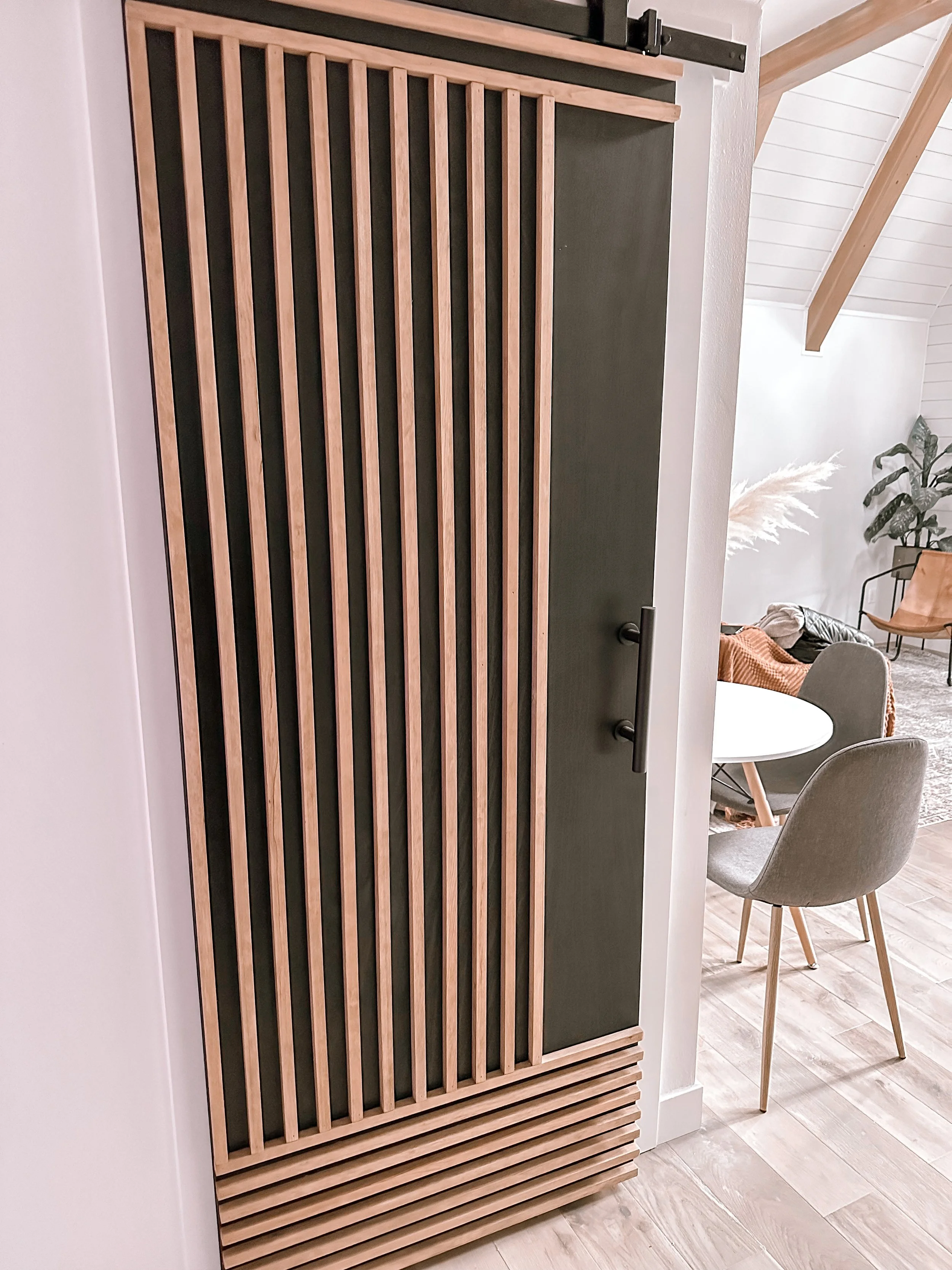 A wooden slatted door with a dark handle, partially open, reveals a room with a round table and beige chairs. The room has white walls, a high ceiling with light-colored wooden beams, and potted plants.