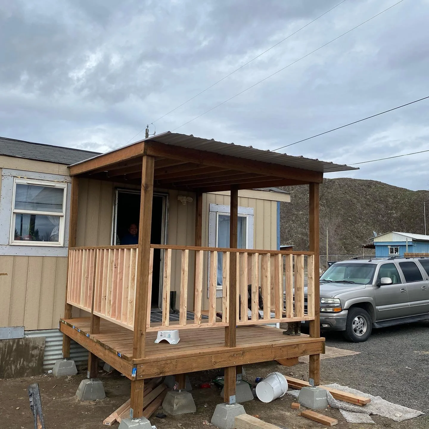 A newly built wooden porch with a roof and railing attached to a beige house, with a person inside and a person partially visible near the stairs. A gray van is parked nearby, and construction materials and debris are visible around the porch on uneven ground.