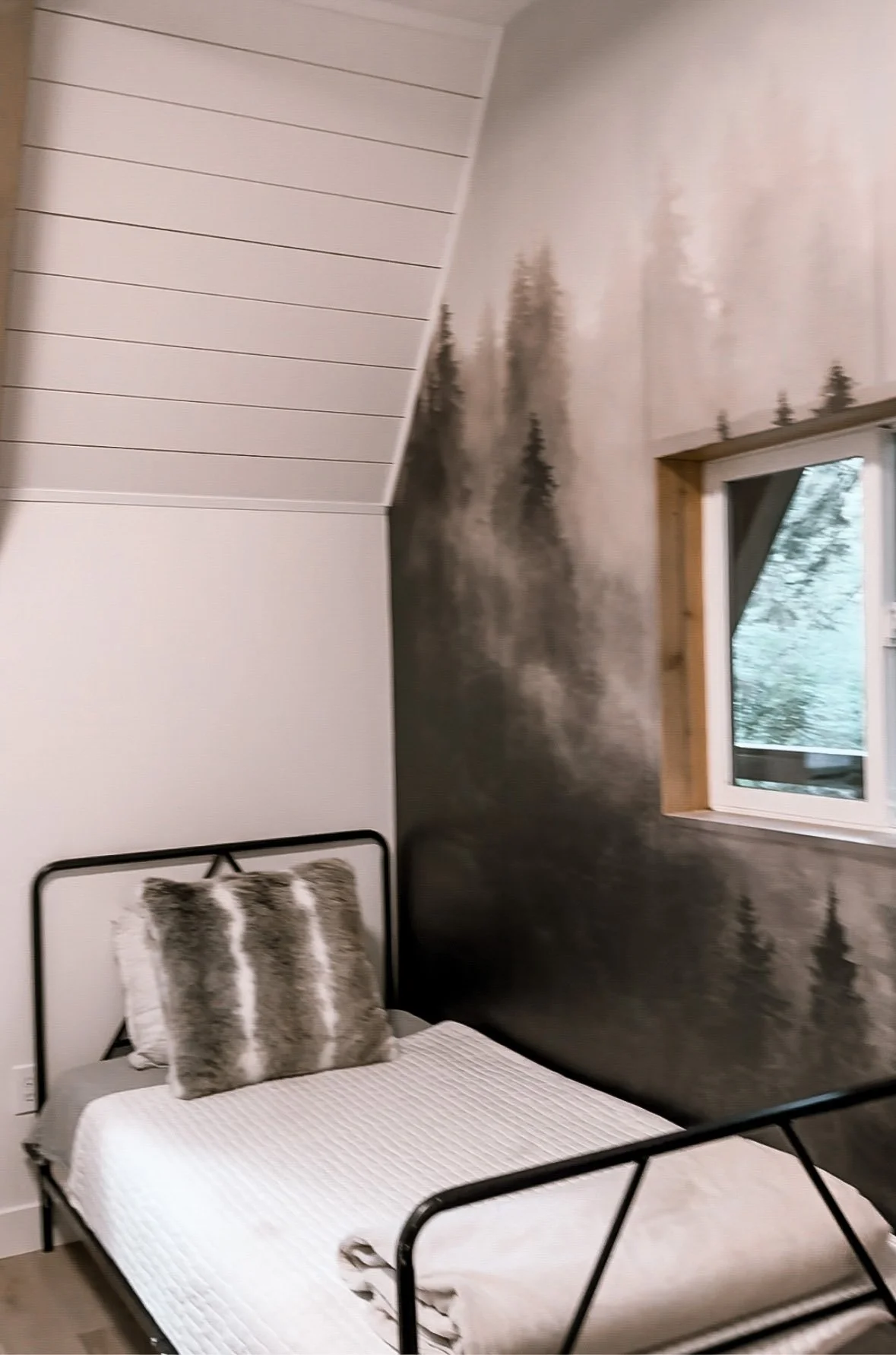 A bedroom with a bed, two faux fur pillows, a painting of a forest on the wall, and a window showing trees outside.