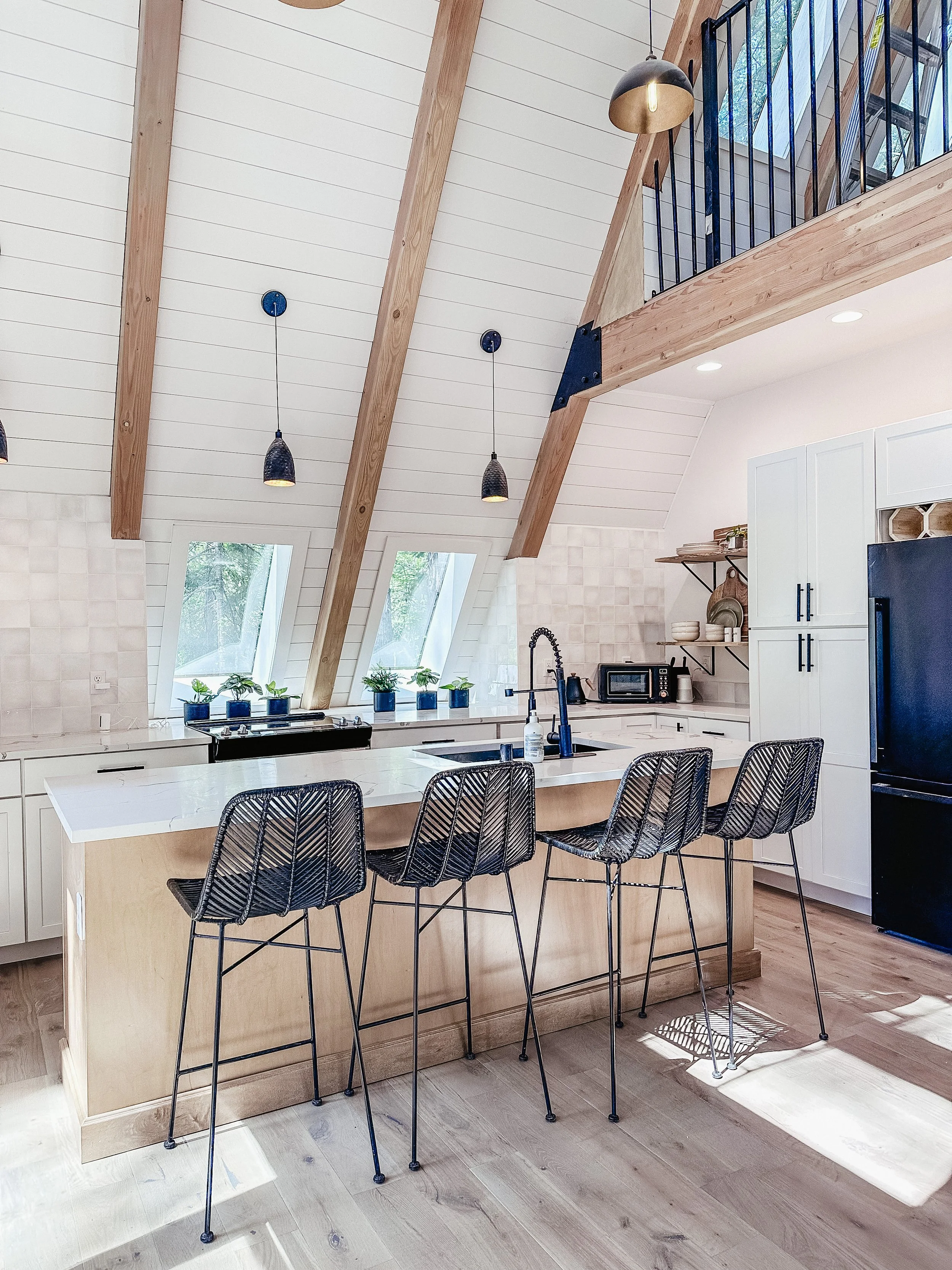 Modern kitchen with white cabinets, black appliances, and a white island counter with four black wire chairs. Sloped ceiling with wooden beams, skylight windows, and hanging pendant lights. Balcony with black railing visible upstairs.