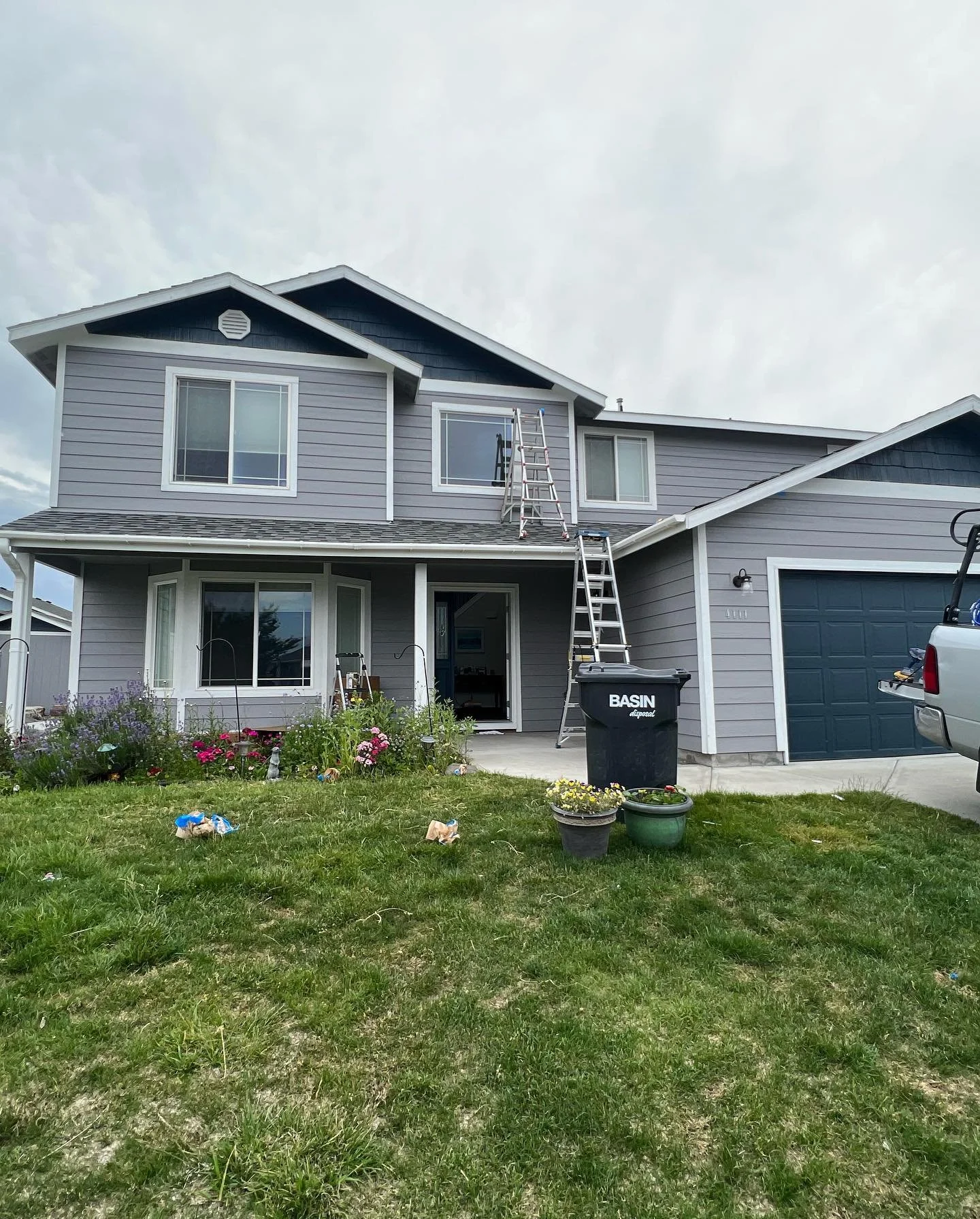 A two-story house with grey siding, white trim, and a dark-colored garage door. There are ladders leaning against the house and a black trash bin in the front yard. The yard has some flowers, plants, and several small garden ornaments scattered across the grass.
