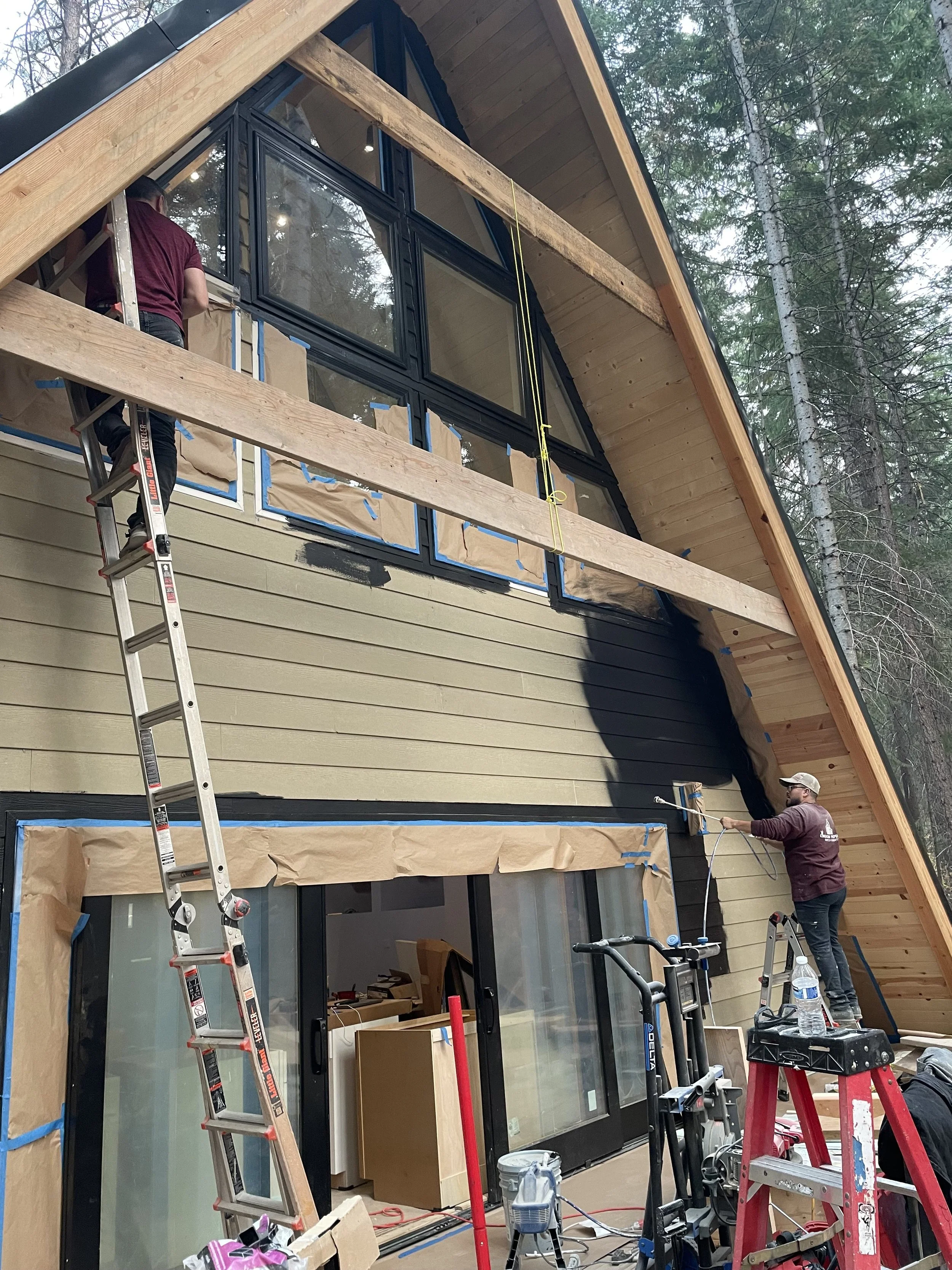 Two workers are installing or working on the exterior wall of a house with dark green siding, large windows, and a wooden roof structure in a wooded area. One worker is on a ladder, and the other is painting or applying coating to the wall.