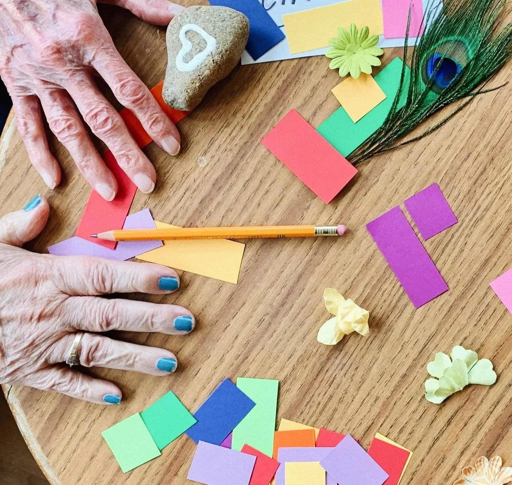 Older hands with blue nail polish arranging coloured paper, a feather and a heart-painted stone