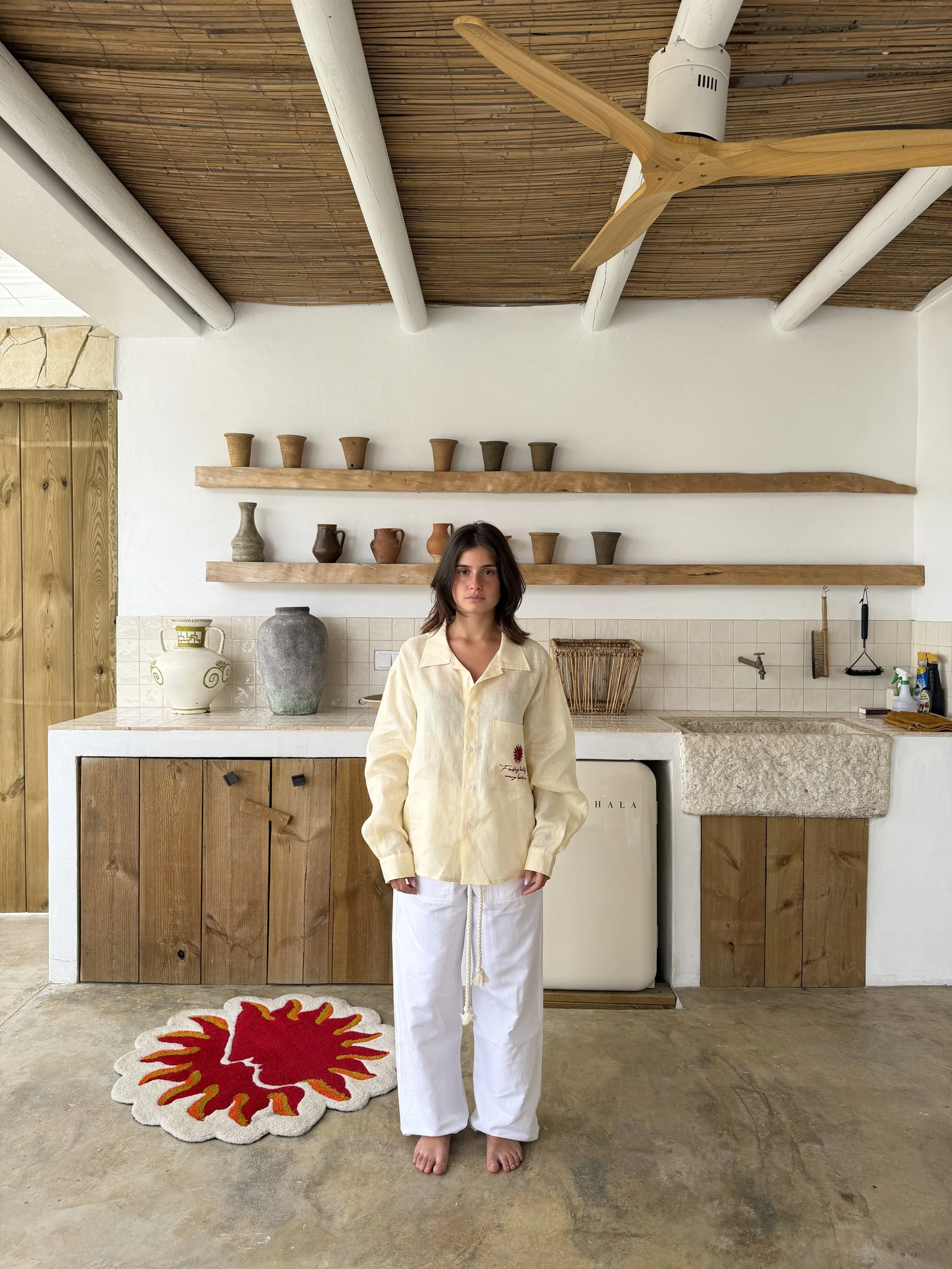A woman standing barefoot on a concrete floor in front of a rustic kitchen setting with wooden shelves holding pottery and decorative objects. She is wearing a cream-colored button-up shirt and white wide-leg pants, with a serious expression, and has shoulder-length dark hair.