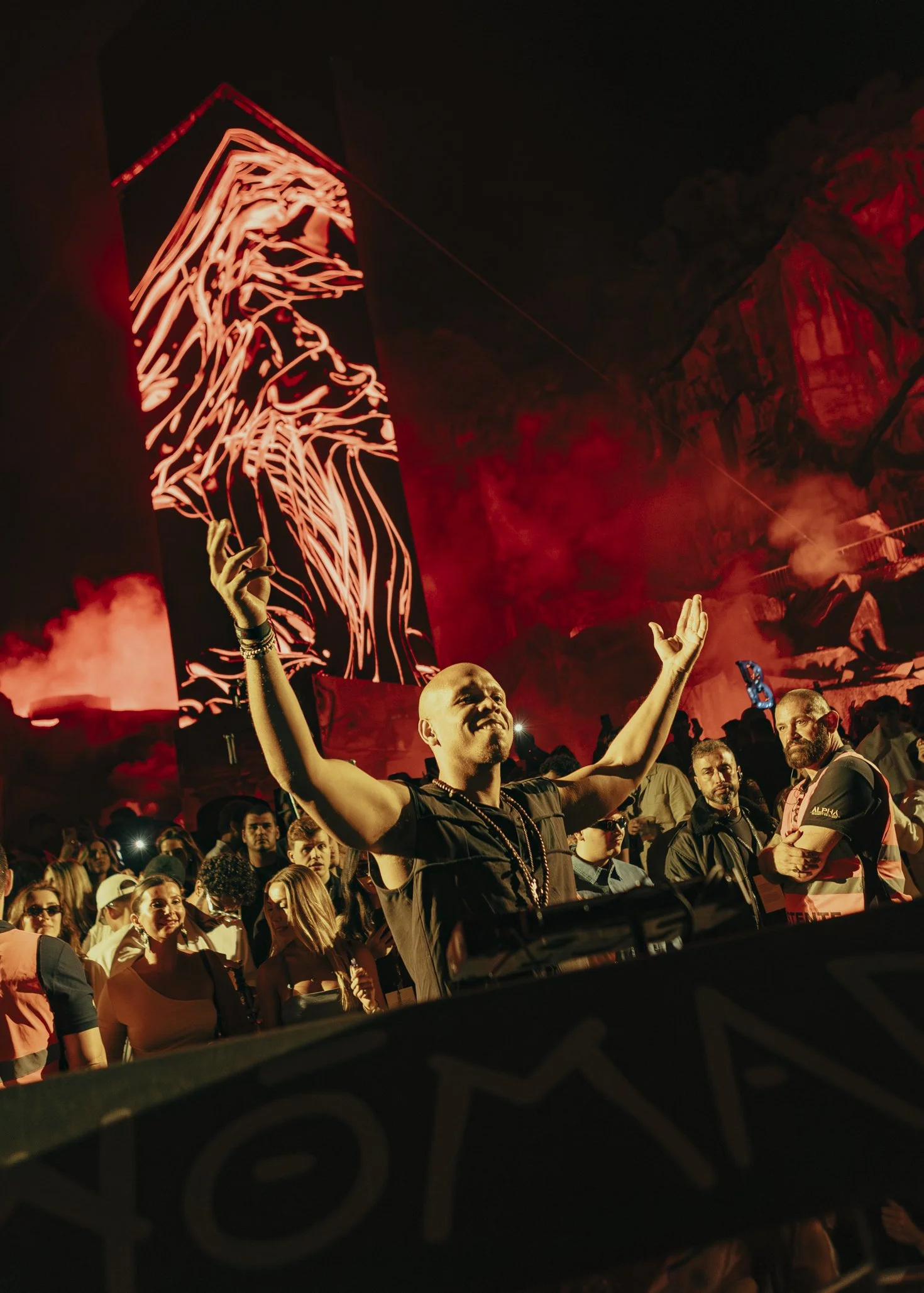 Francis Mercier DJ at a concert or festival, surrounded by a crowd of people, with a large screen displaying red and black abstract art in the background.