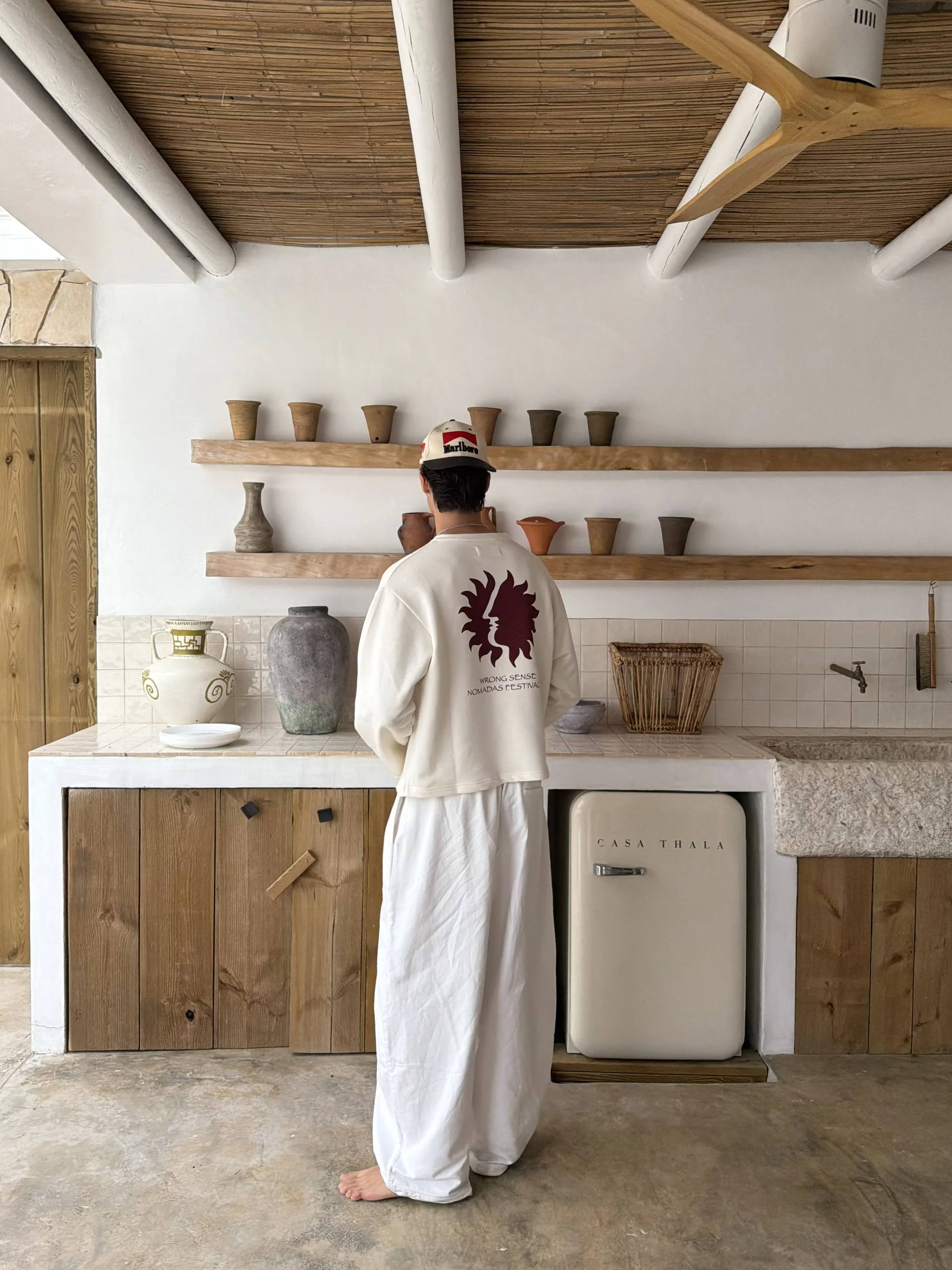 Person with short dark hair wearing a white cap with red and black logos, a long white robe with a red lion emblem on the back, standing barefoot in front of shelves with various antique or decorative pottery, in a room with wood paneling on the ceiling and a white wall.