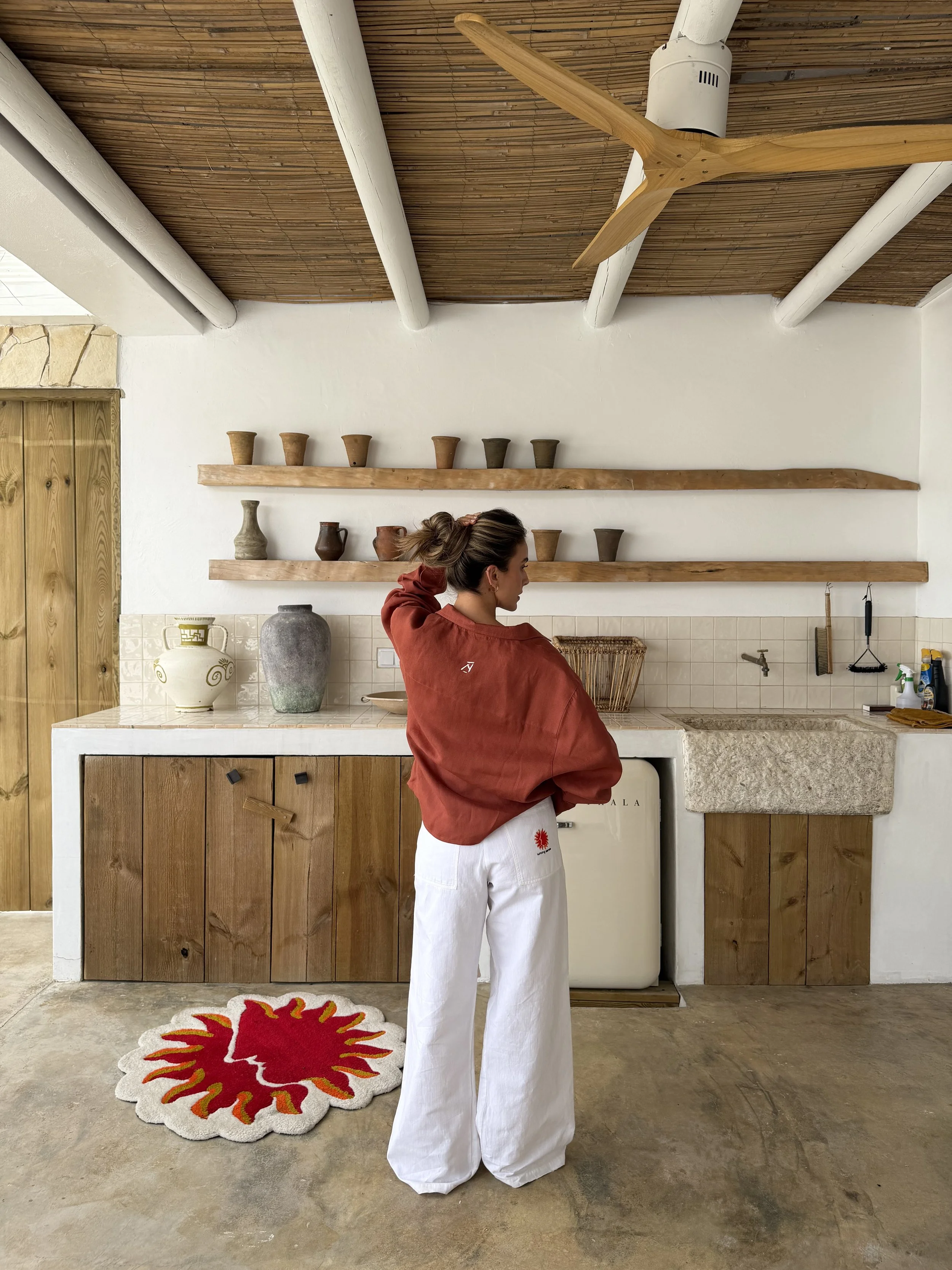 A woman standing indoors in front of a wooden cabinet and white wall with two wooden shelves holding pottery. She is wearing white wide-leg pants and a red jacket, with her hair tied up. A colorful sun-shaped rug with a face is on the floor in front of her.