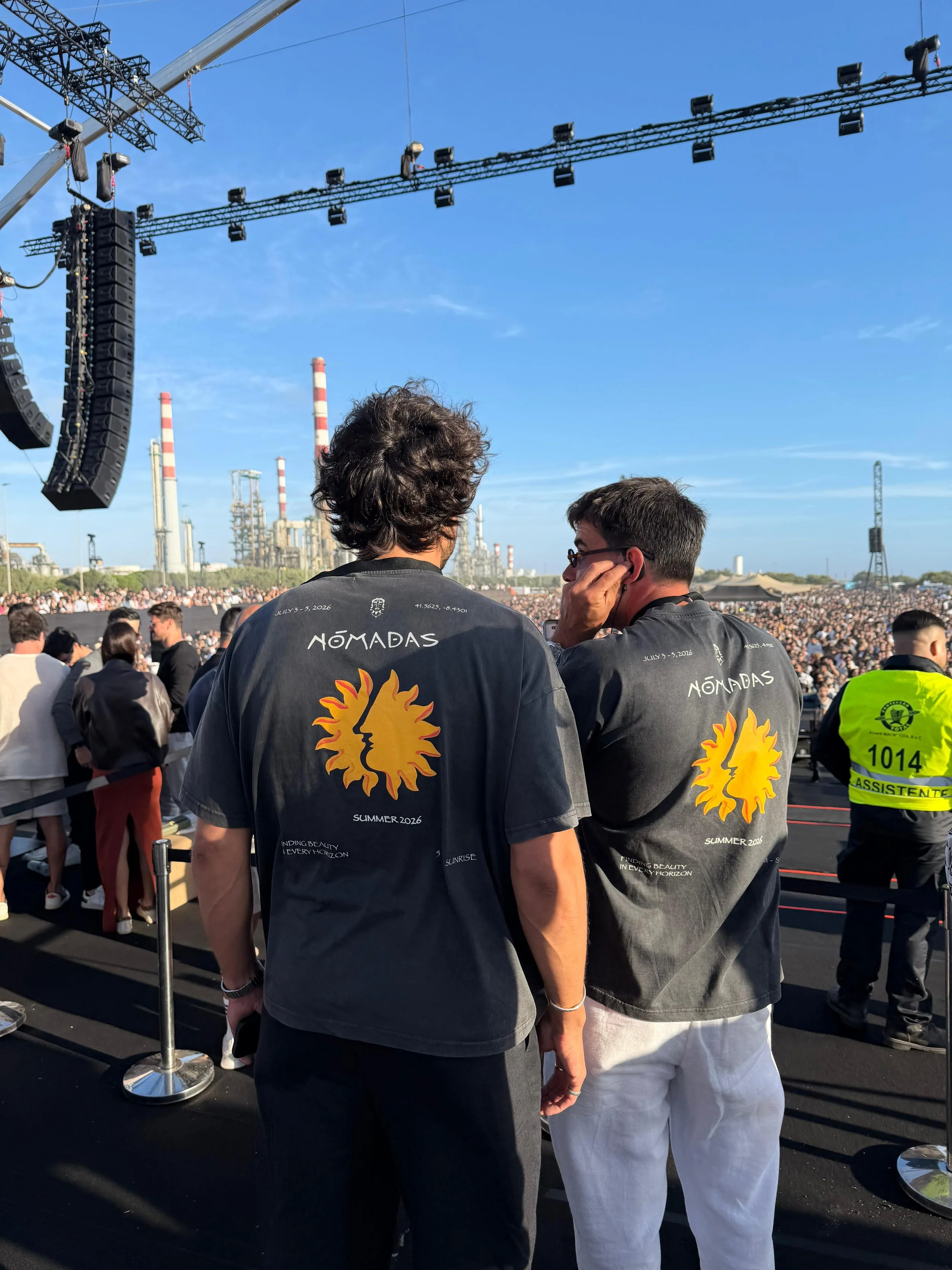 Two men wearing black jackets with a sun and face design on the back, standing in a crowd at an outdoor event, with industrial buildings and blue sky in the background.