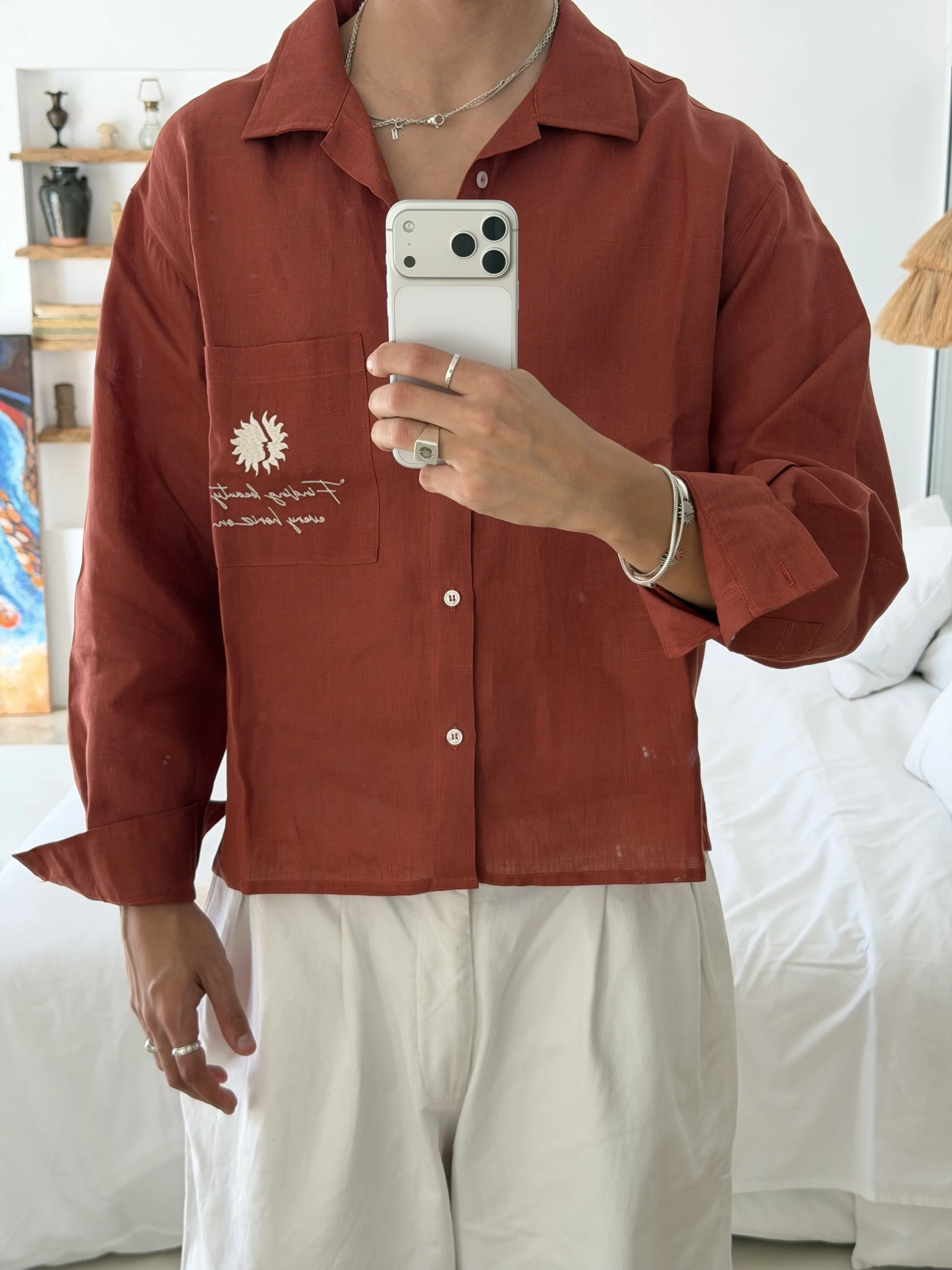 Person wearing a red shirt with embroidery, taking a mirror selfie in a room with shelves and decorative objects.