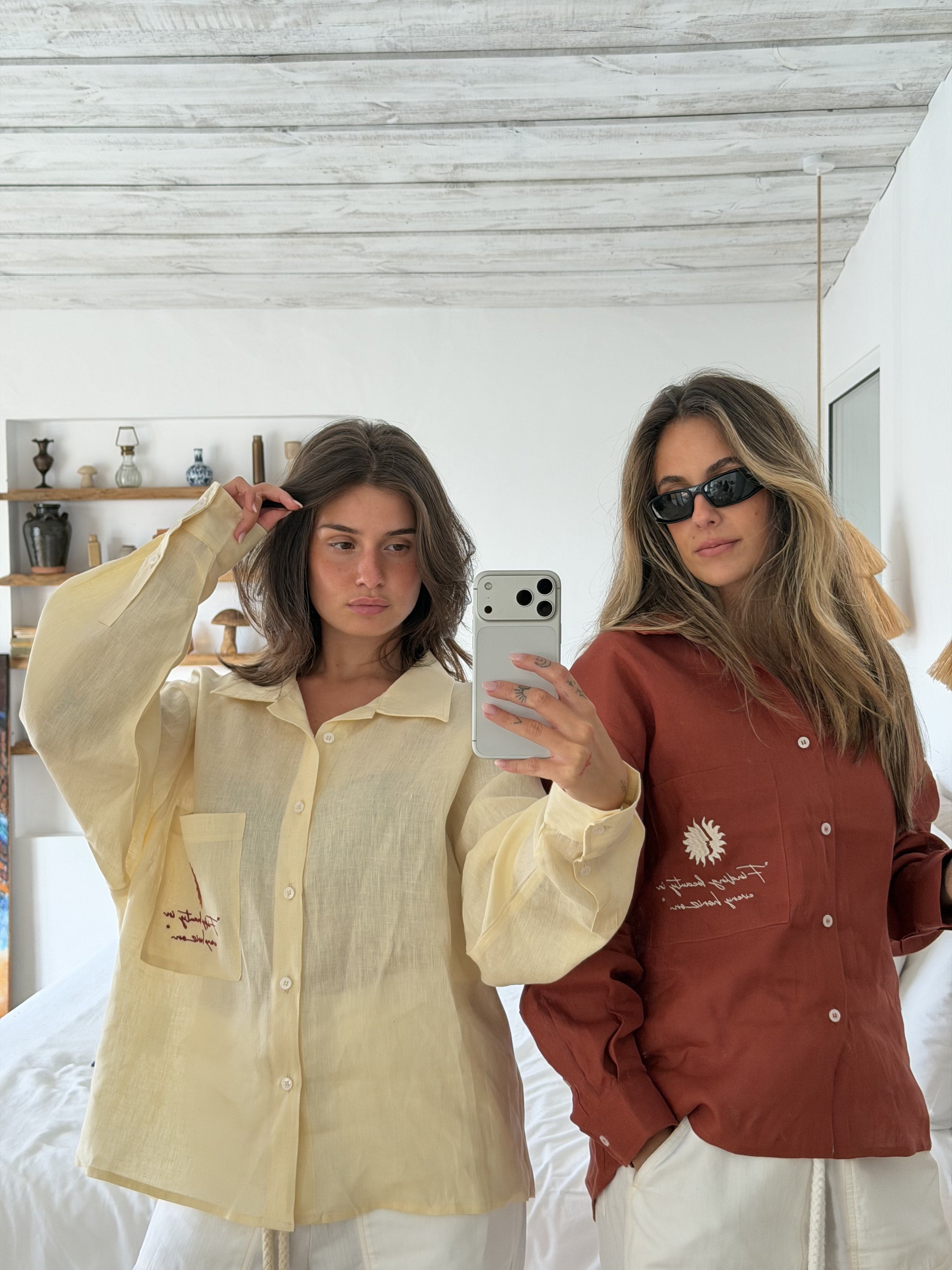 Two women are taking a mirror selfie. One woman has wavy light brown hair, wears black sunglasses, a brownish-red shirt with writing, and white pants. The other woman has straight dark brown hair, wears a yellow shirt with writing, and white pants. They are inside a room with white walls, a wooden shelf with decorative items, and a white bed in the background.