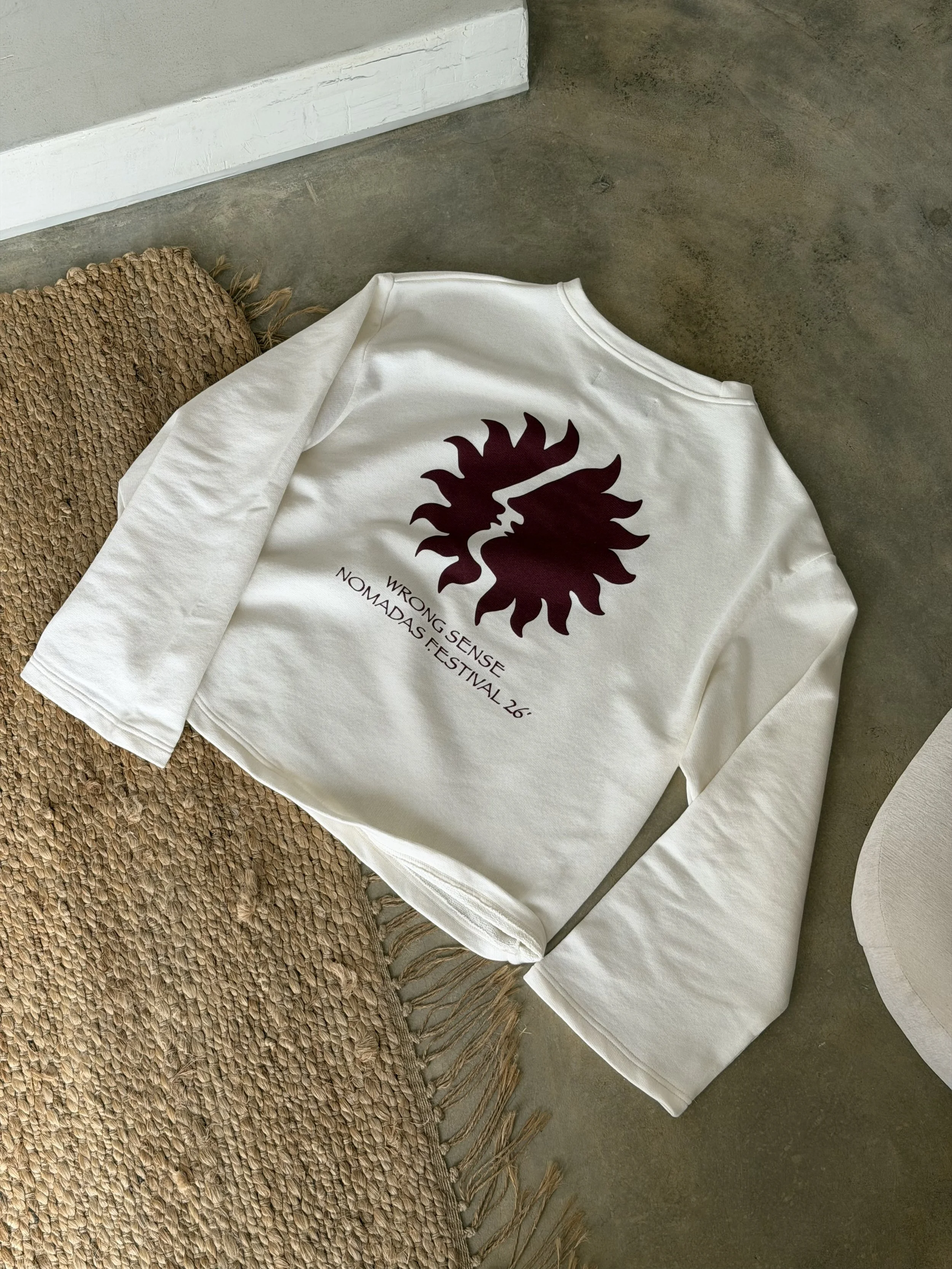 A white sweatshirt with a dark red lion logo and text, laid on a textured brown rug on a concrete floor, near a white baseboard and a white chair.