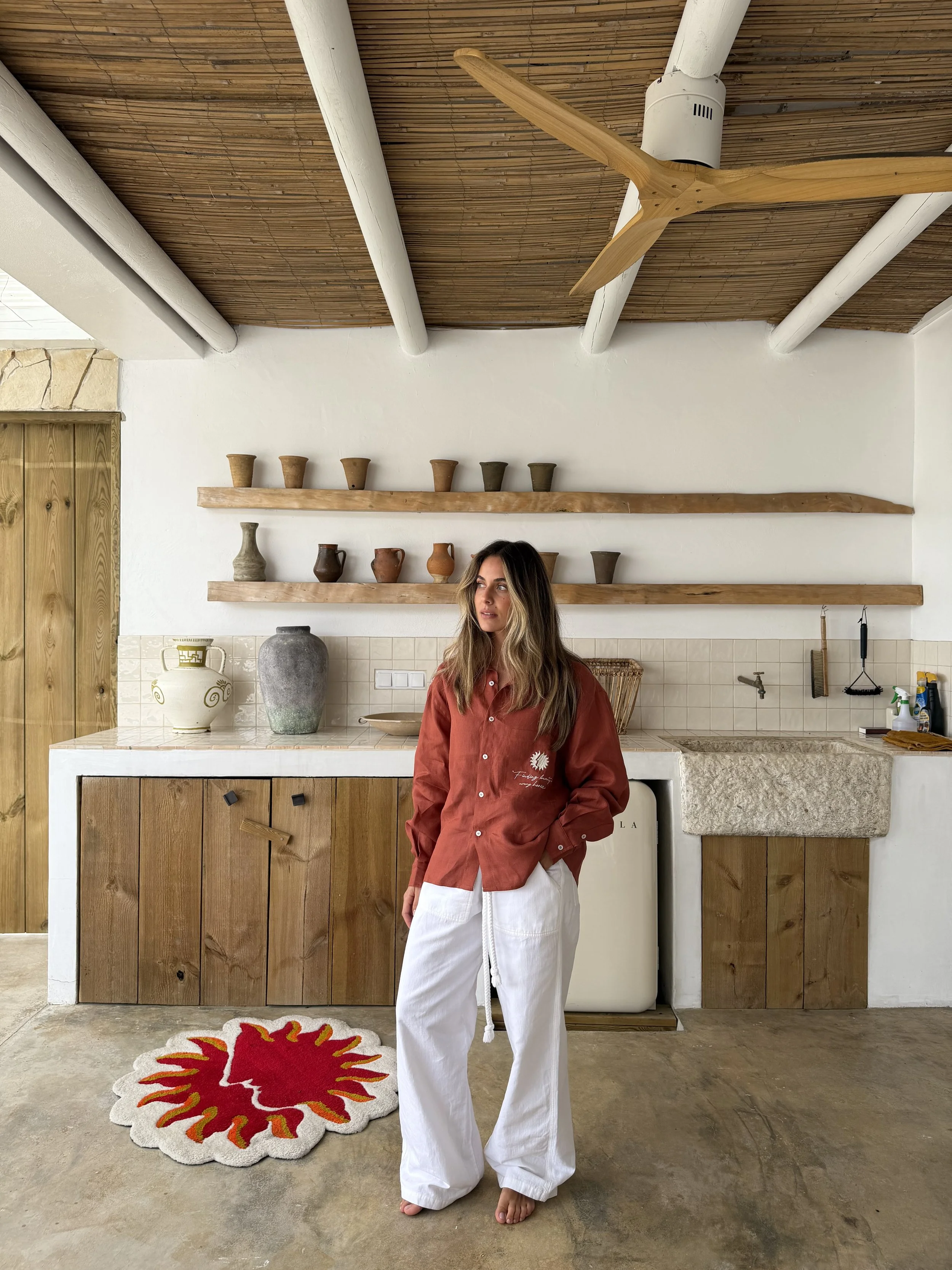 A woman standing barefoot in a kitchen with a rustic and modern decor, wearing a red button-up shirt and white wide-leg pants, in front of a whitewashed wall with wooden shelves holding pottery and decorative items, a ceiling fan, and a colorful sun-shaped rug on the floor.