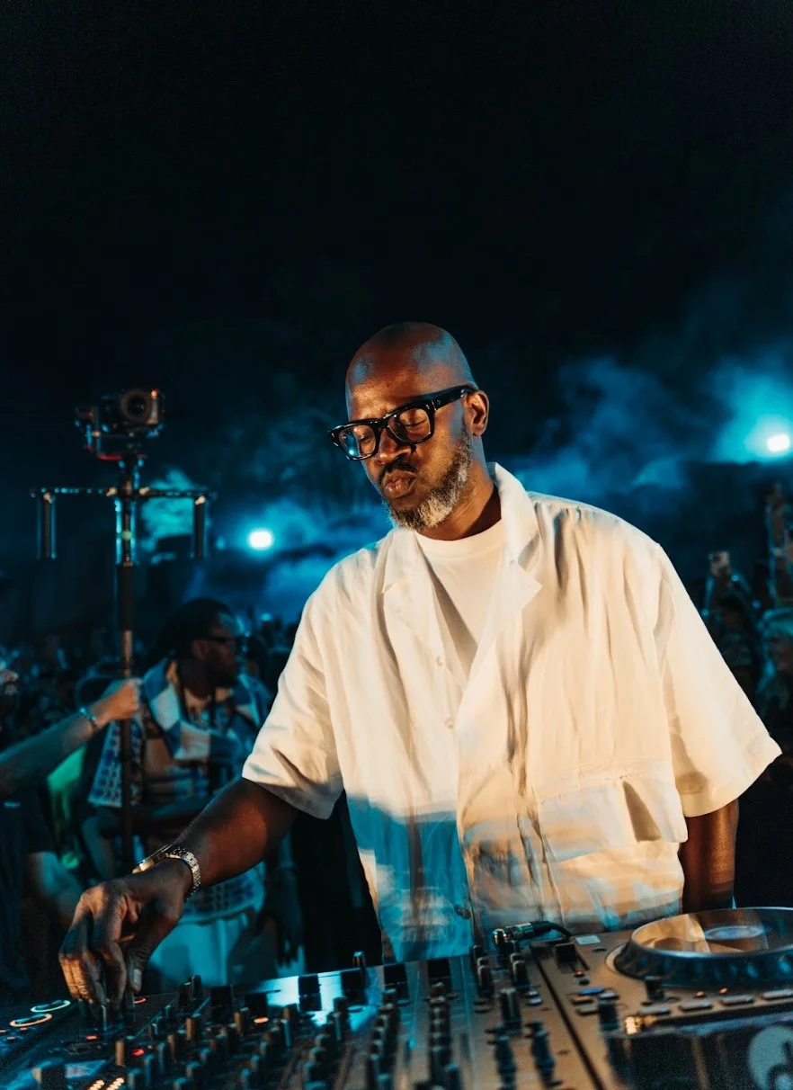 Black Coffee mixes music at a nighttime outdoor event, with blue lighting and a crowd in the background.