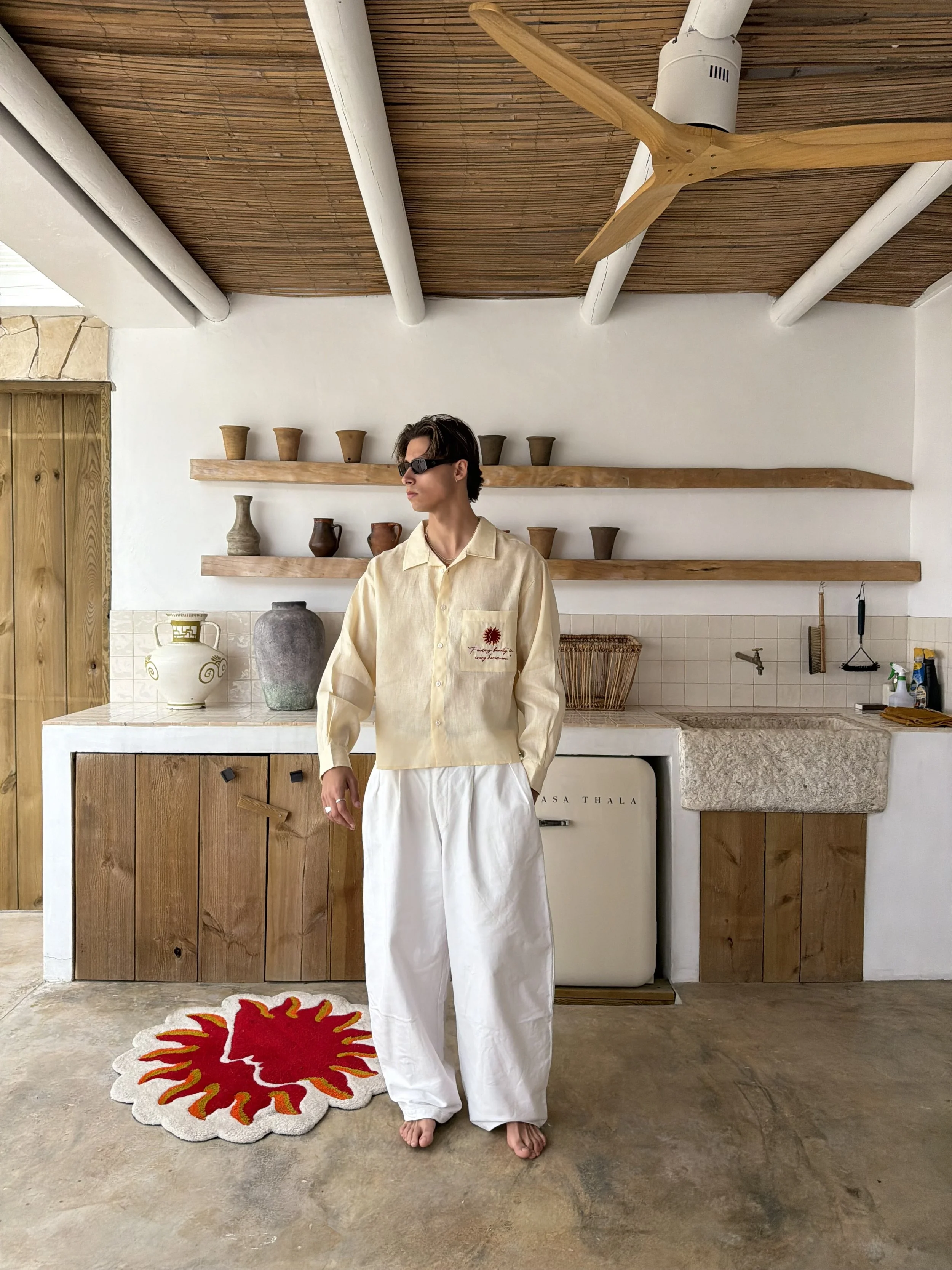 A person standing in a kitchen with a rustic, modern aesthetic, wearing sunglasses, a light yellow shirt with a red logo, and white wide-legged pants. The kitchen features wooden cabinets, white tiled walls, decorative pottery on shelves, a red and white floral rug, and a wooden ceiling with exposed beams and a ceiling fan.