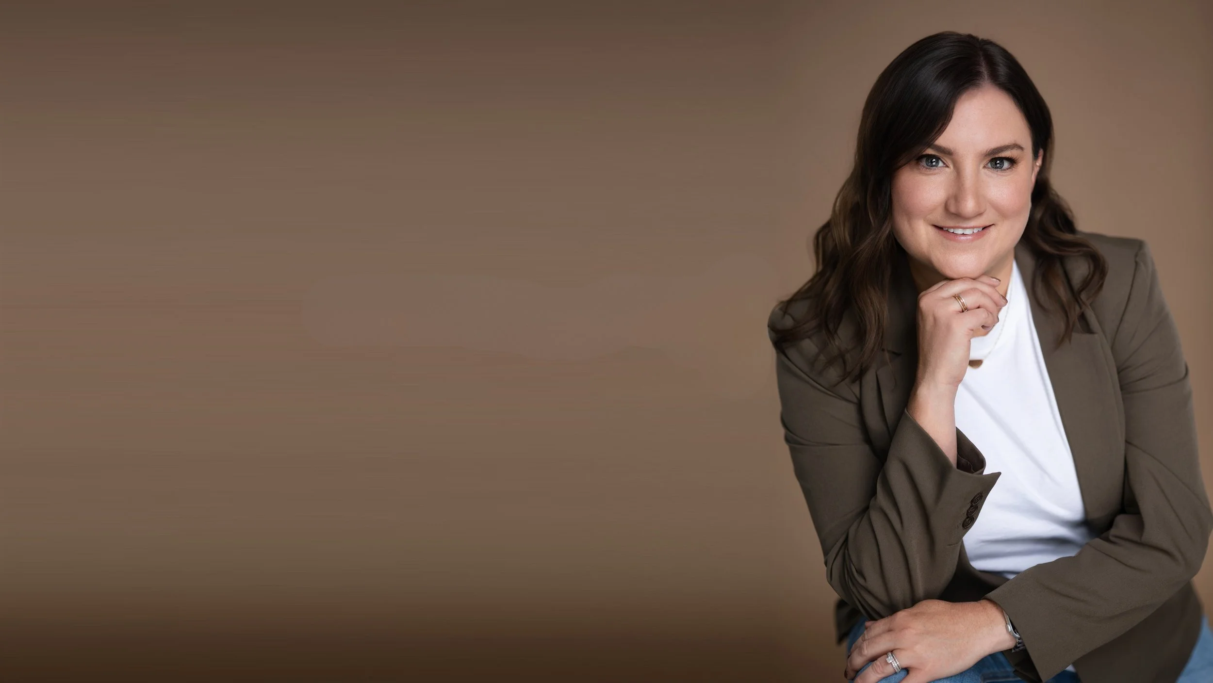 A woman with long brown hair, wearing a tan blazer and white top, smiling and resting her chin on her hand, sitting against a plain brown background.