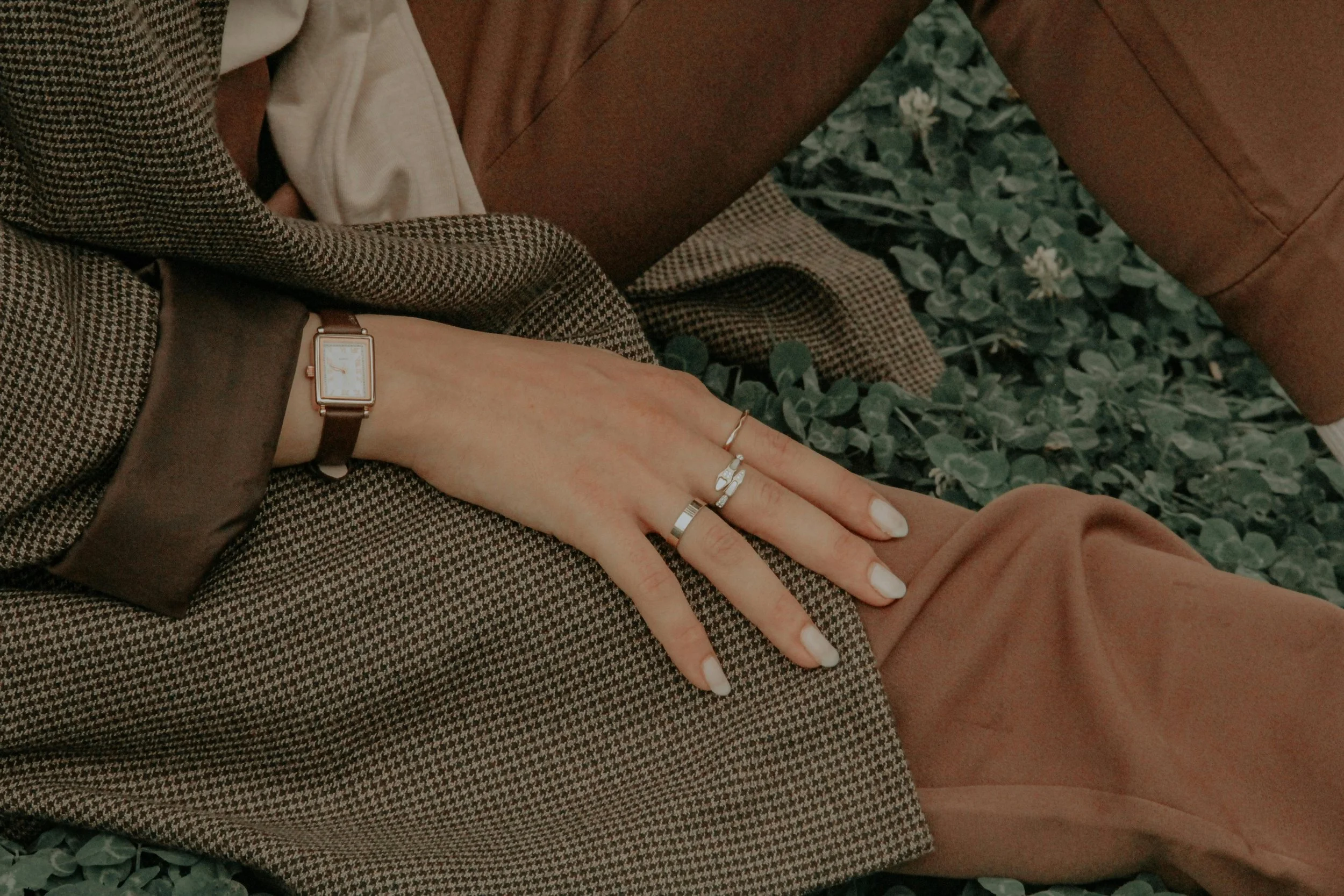 Close-up of a woman's hand resting on her lap, wearing a brown checkered blazer, a watch, and multiple rings, against a backdrop of green foliage.