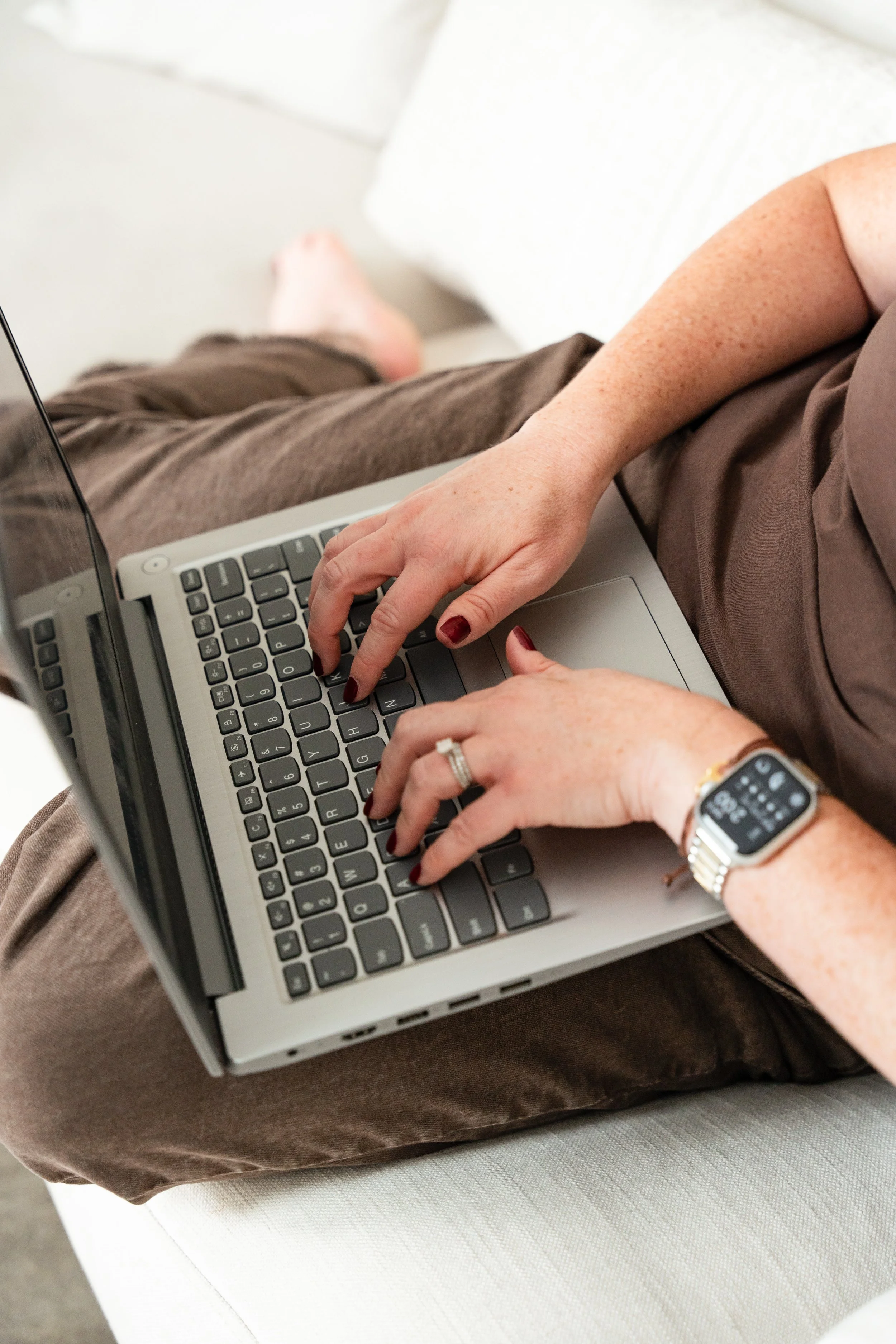 A person typing on a laptop keyboard with a smartwatch on their wrist, sitting on a cushioned surface