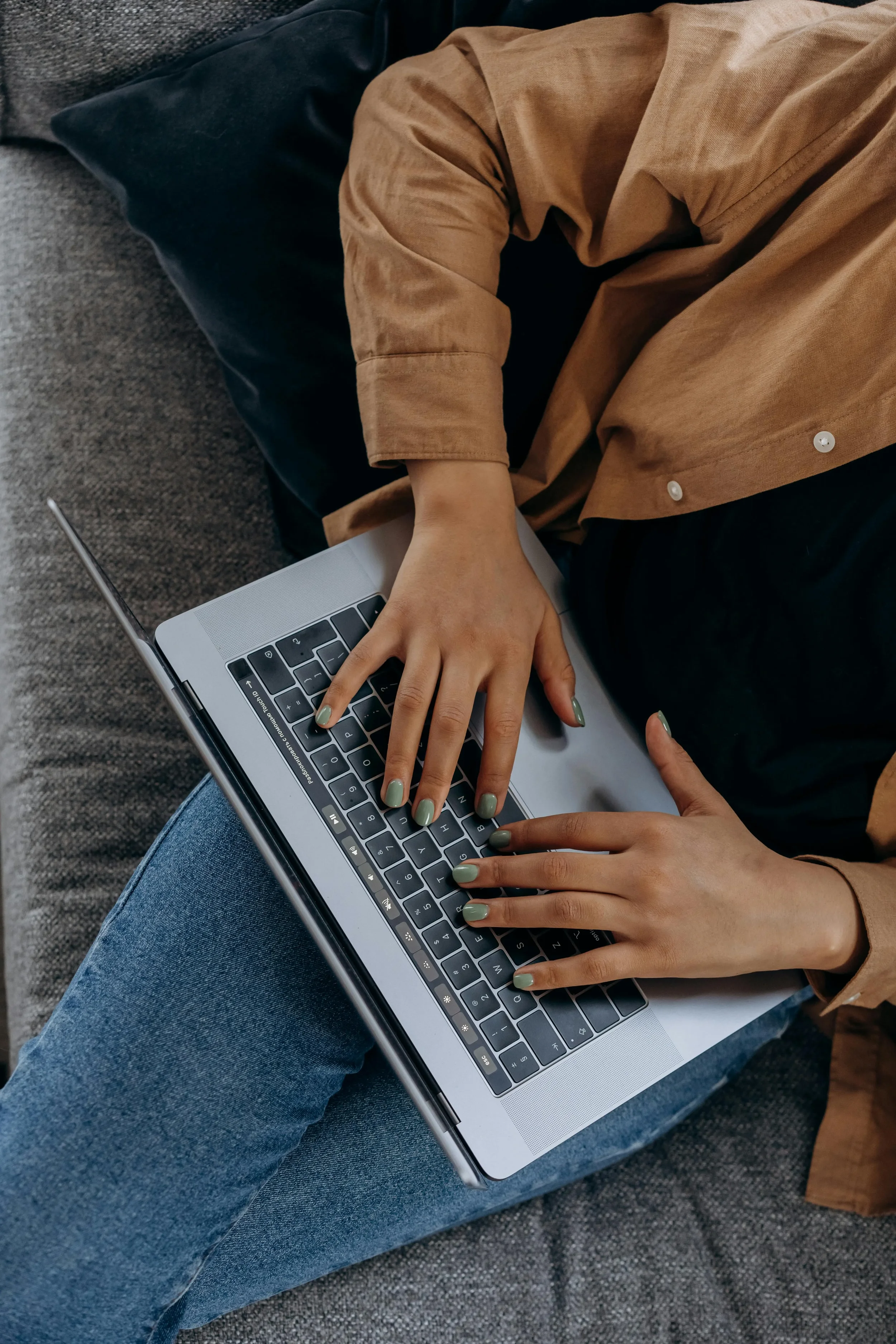 Person using a laptop on their lap while sitting on a gray couch, wearing blue jeans and a tan long-sleeve shirt.