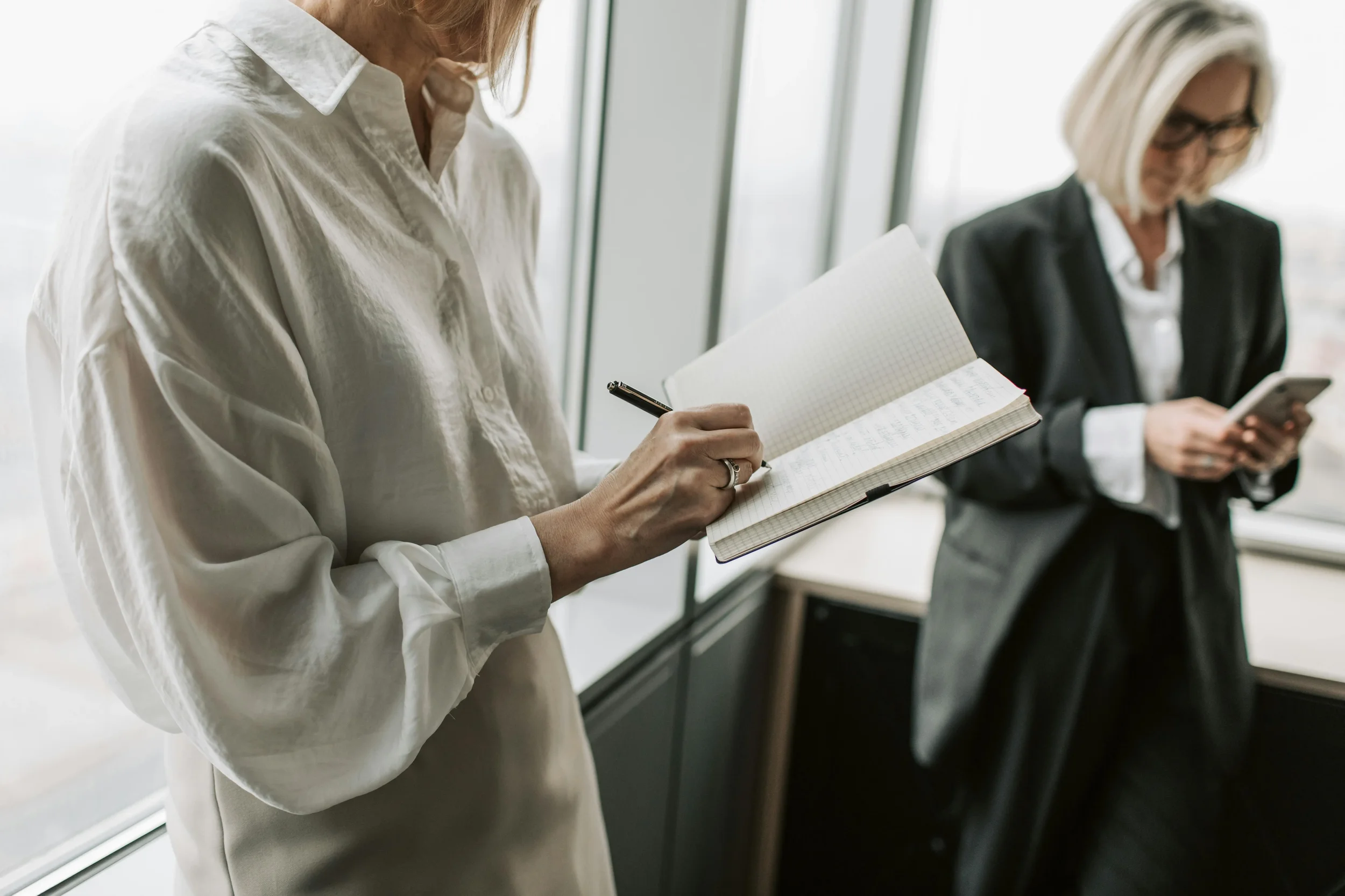 Two women in business attire are standing near a large window in an office, with one holding a notebook and pen, and the other looking at their phone.