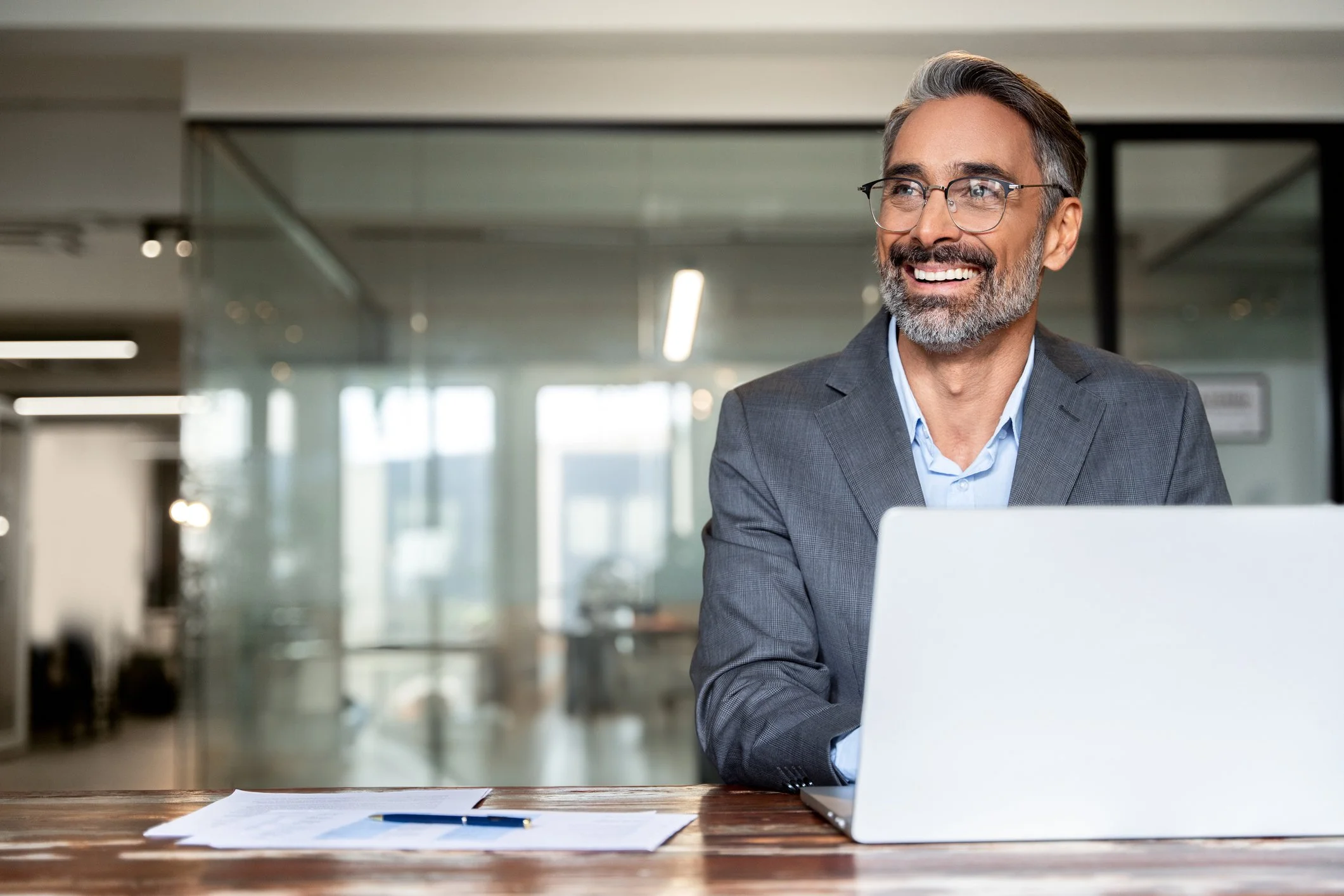 A smiling man with glasses and gray hair sitting at a desk with a laptop in a modern office setting.
