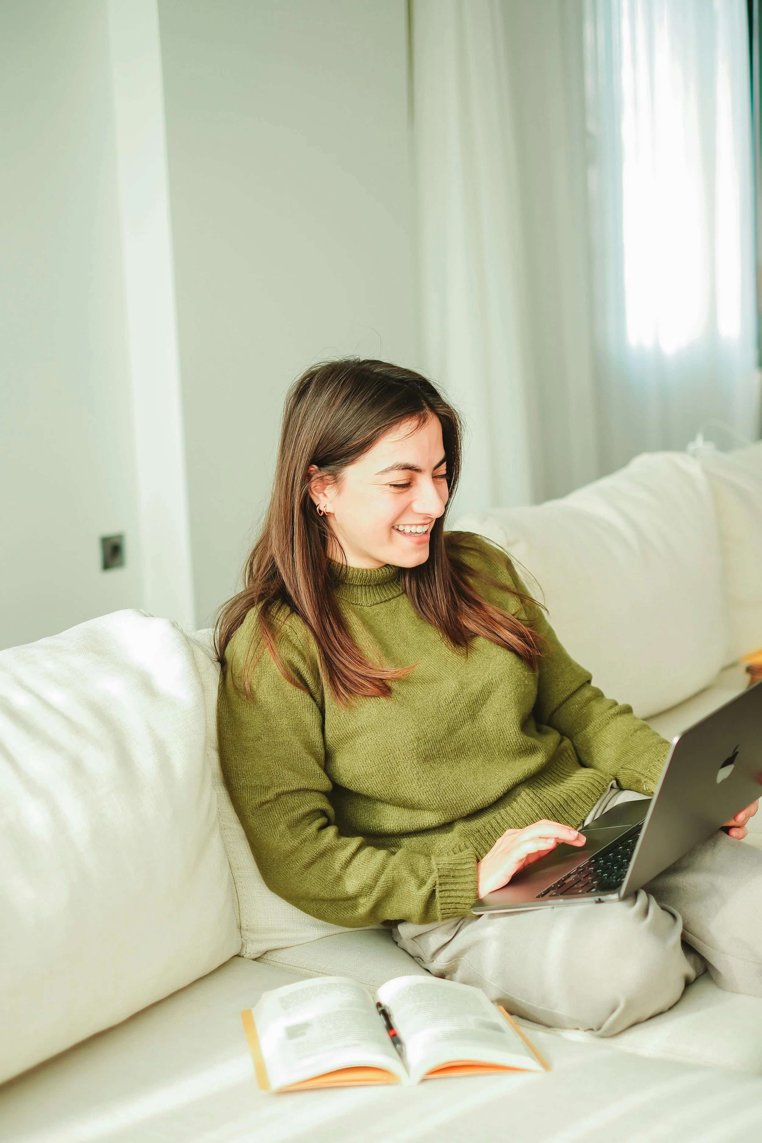 A young woman in a green sweater sitting on a white sofa, using a laptop, with an open book beside her, in a bright room with light-colored walls and windows.