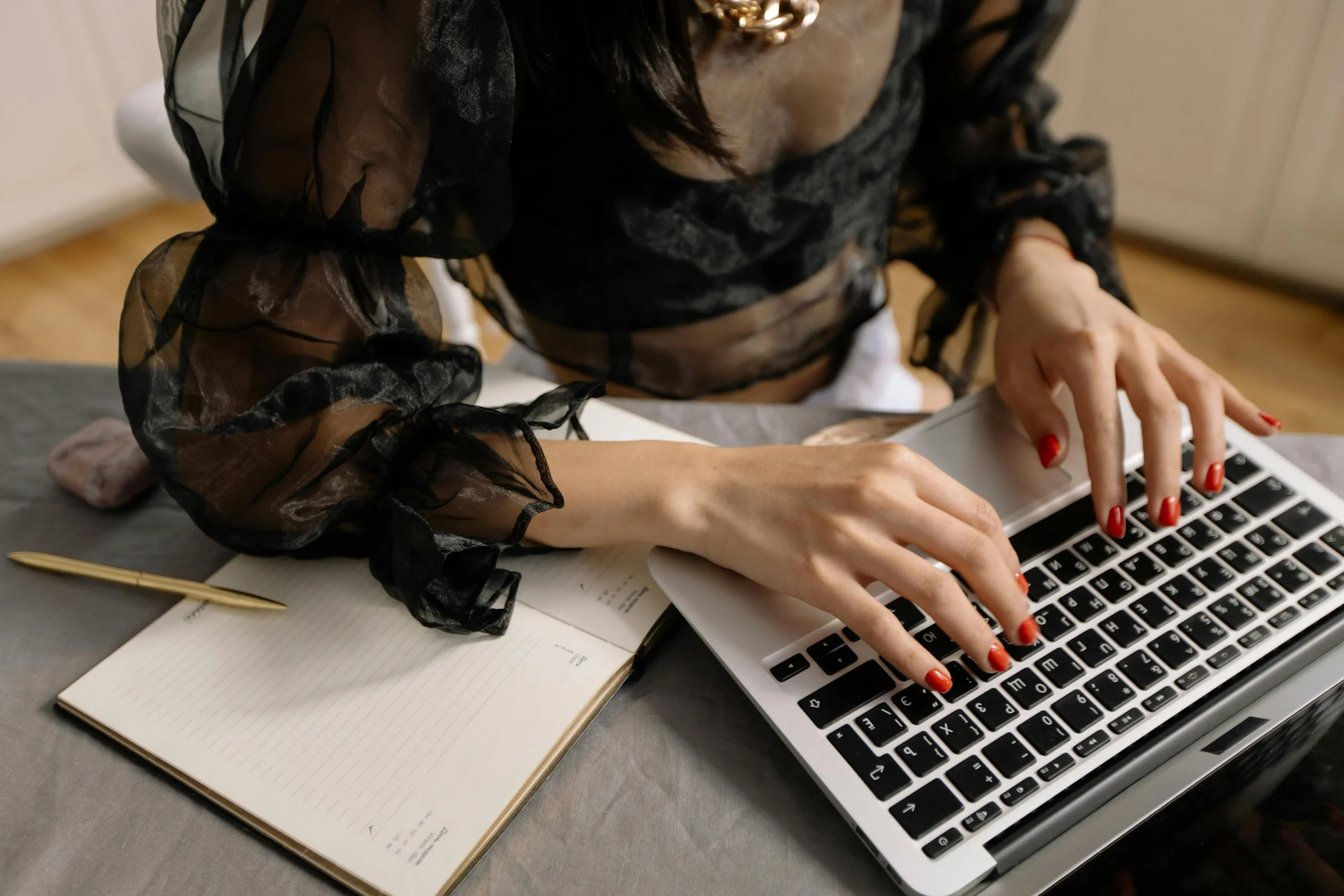 A person typing on a laptop keyboard on a desk with a notepad, a gold pen, and a pink object. The person is wearing a black sheer blouse with ruffled sleeves and has red nail polish.