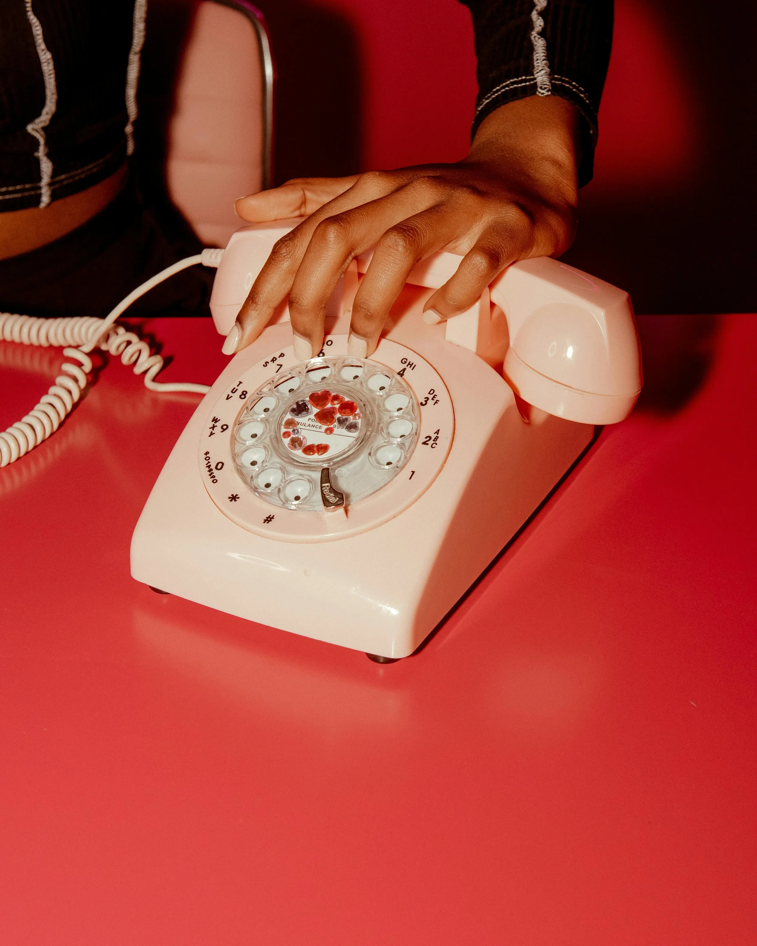 A person's hand dialing a rotary phone with a clear center showing red and black beads on a pink surface.