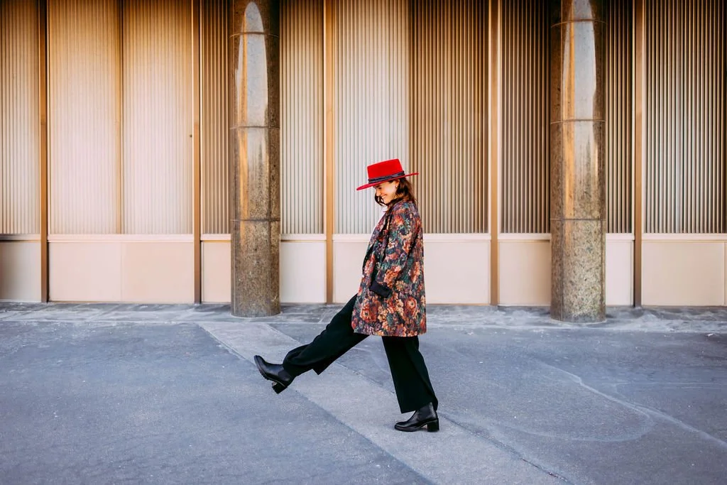 Woman in a floral patterned coat, black pants, and black shoes, wearing a wide-brimmed red hat, standing on a grey pavement in front of a beige wall with vertical panels and stone columns, kicking her leg in a playful pose.