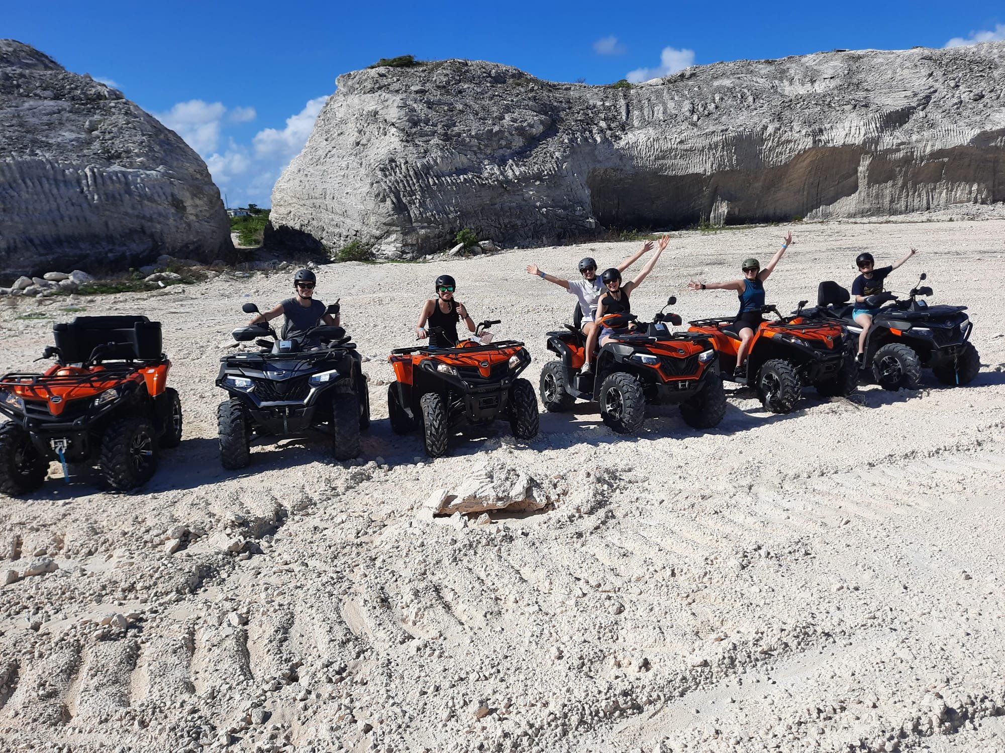 Group of six people wearing helmets riding all-terrain vehicles (ATVs) on sandy terrain with large rocks and cliffs in the background,  J & S Caribbean Rentals, Grand Turk Island.