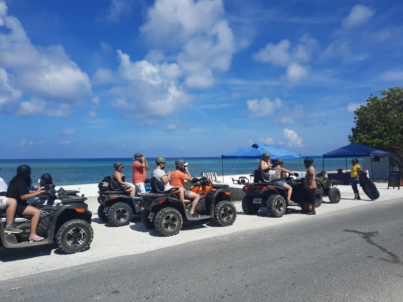 Group of people on all-terrain vehicles (ATVs) near the beach with ocean and blue sky in the background,  J & S Caribbean Rentals, Grand Turk Island, and tents set up nearby.