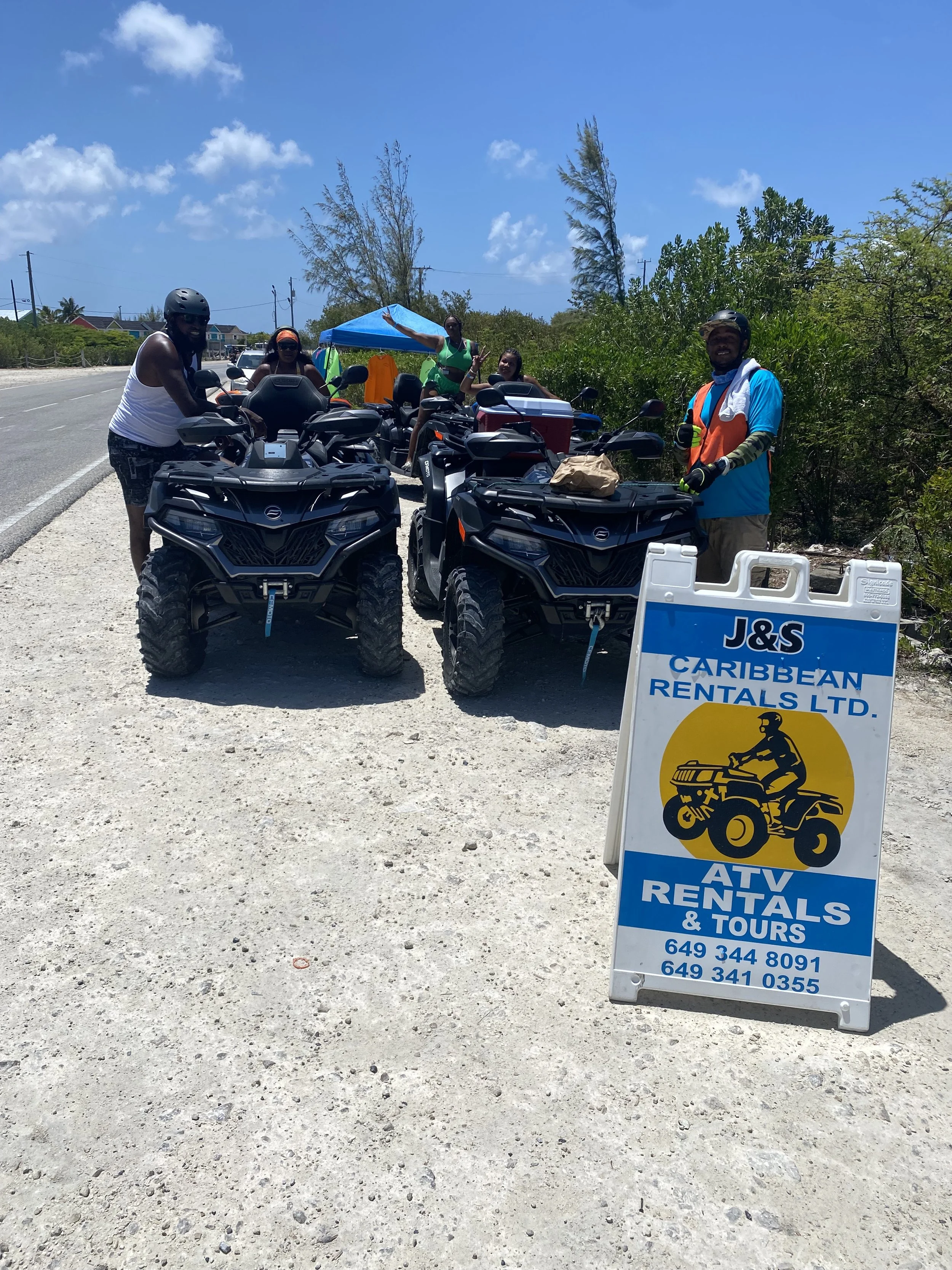 Group of people with ATVs and UTVs parked on the side of a road, with a sign for J&S Caribbean Rentals LTD for ATV rentals and tours Grand Turk Island.