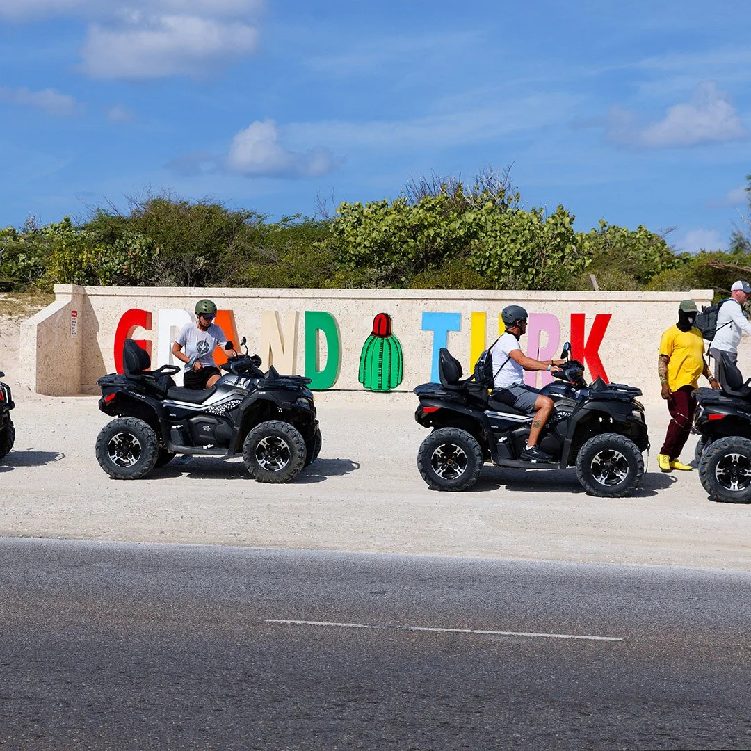 People riding all-terrain vehicles (ATVs)  J & S Caribbean Rentals, Grand Turk Island on a roadside with a colorful sign that spells out 'Grand Turk' and features a cactus graphic, and a background of blue sky and green bushes.